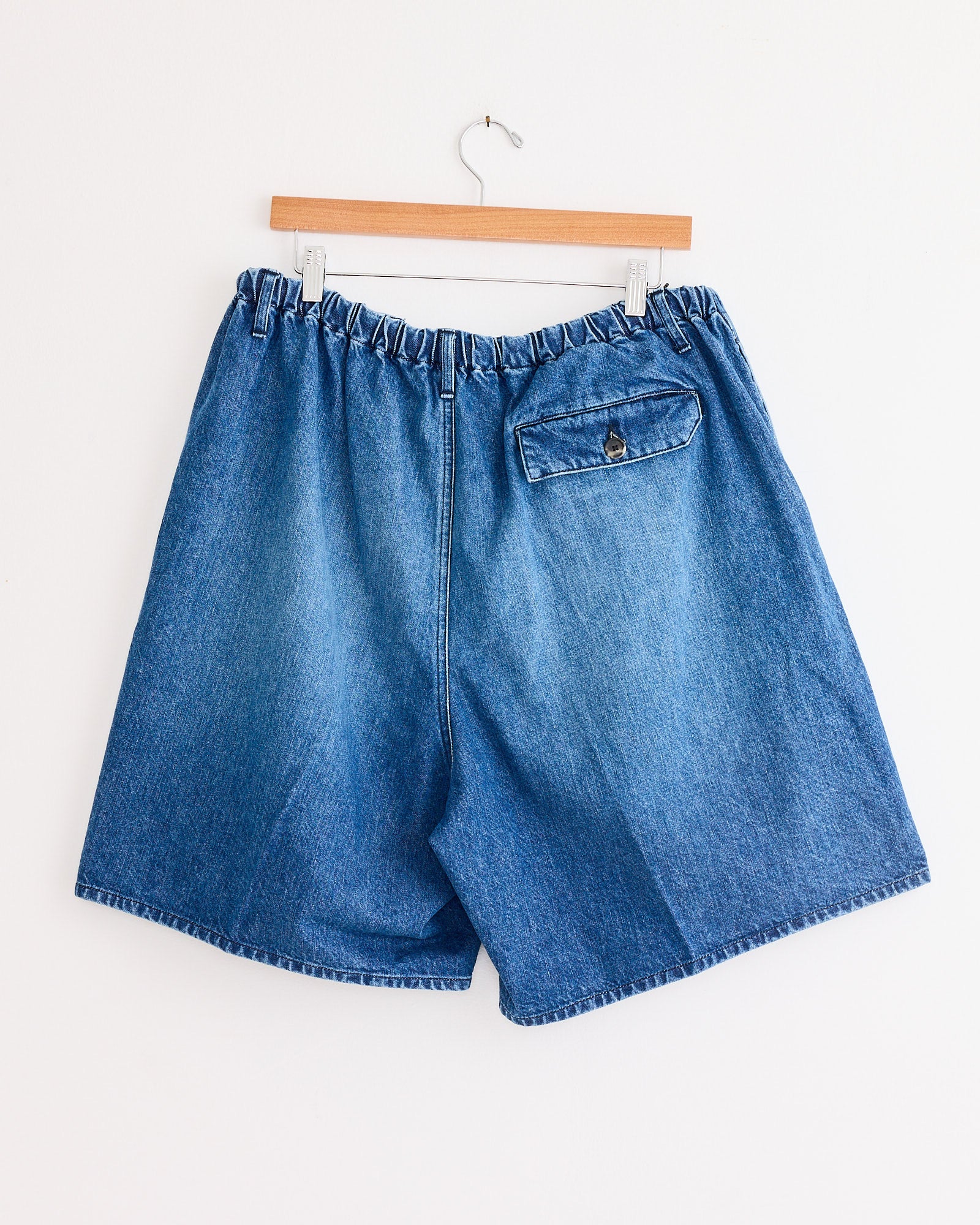 The Sillage Circular Fatigue Short in Blue Denim Damaged features an elastic waistband and a single buttoned back flap pocket, shown on a wooden hanger against a white wall.