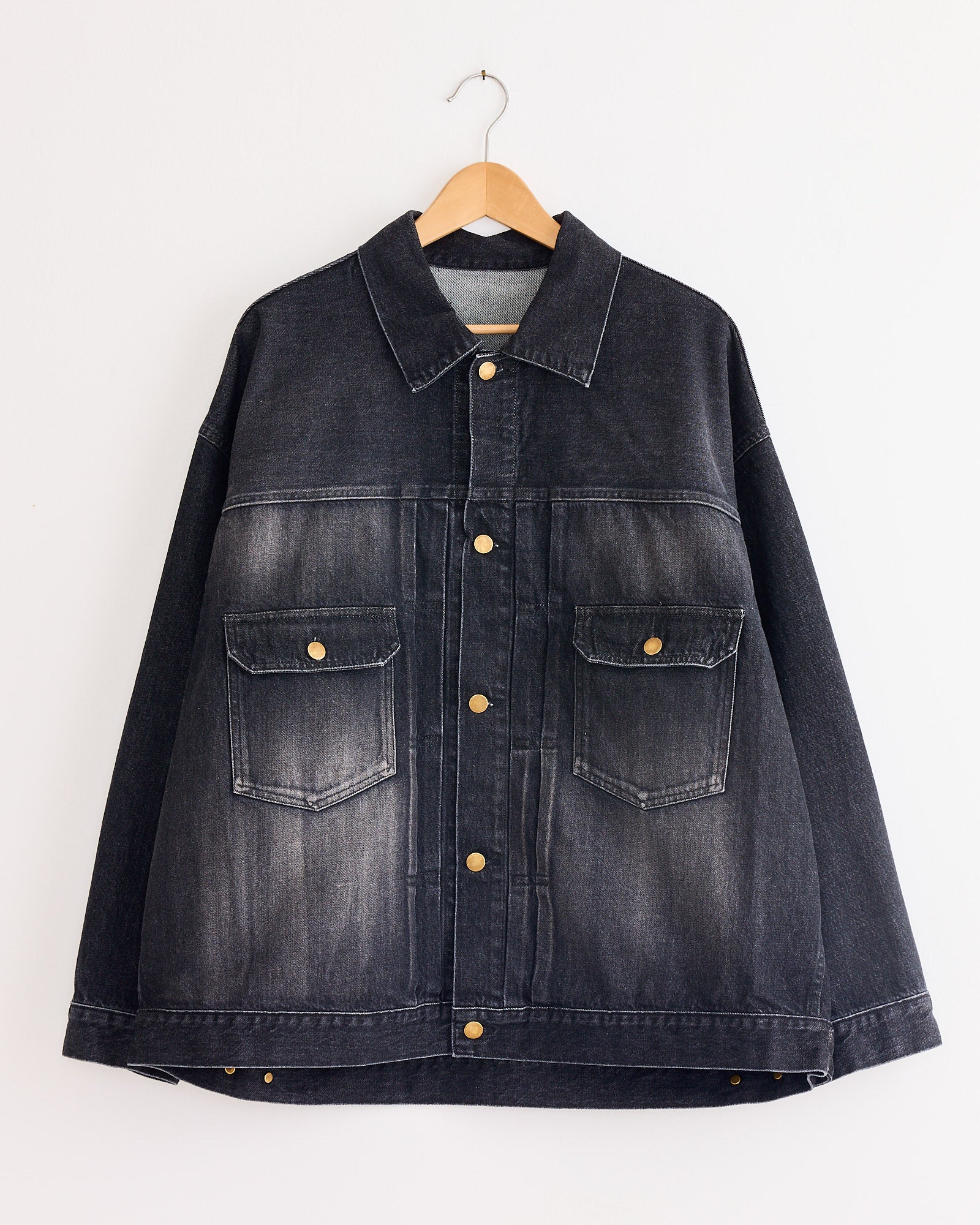 The Sillage Trucker Jacket in Black Denim Damage, featuring brass buttons and two chest pockets, hangs on a wooden hanger against a white background.