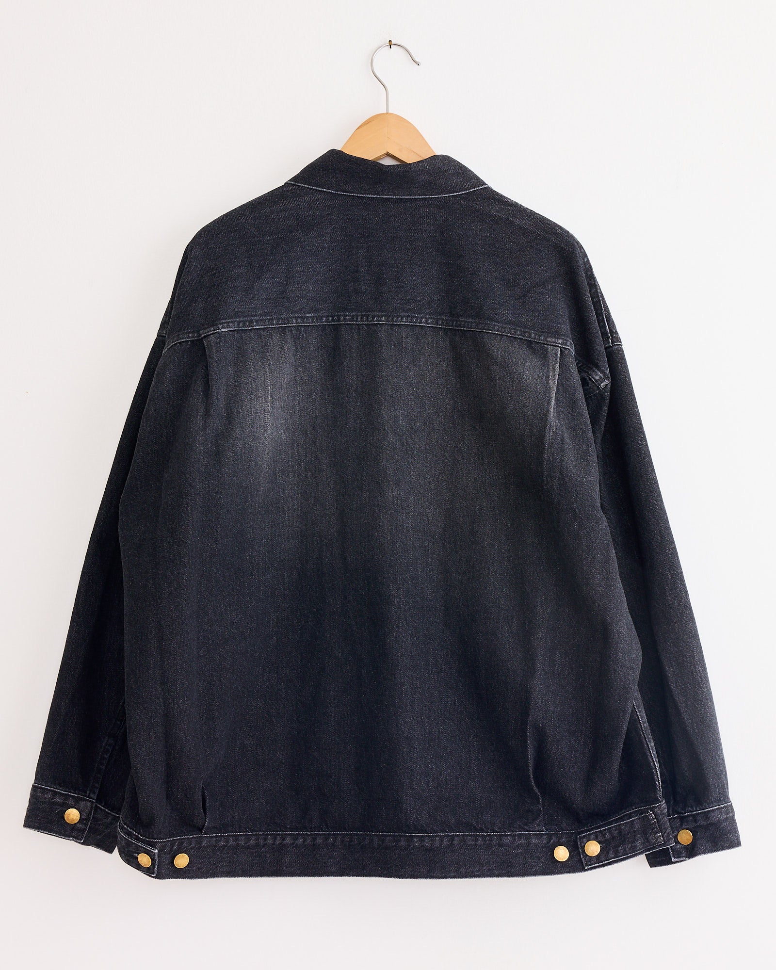 The Sillage Trucker Jacket in Black Denim Damage, crafted in Japan, hangs on a wooden hanger against a white wall, displayed from the back with gold buttons accenting the cuffs and waist tabs.