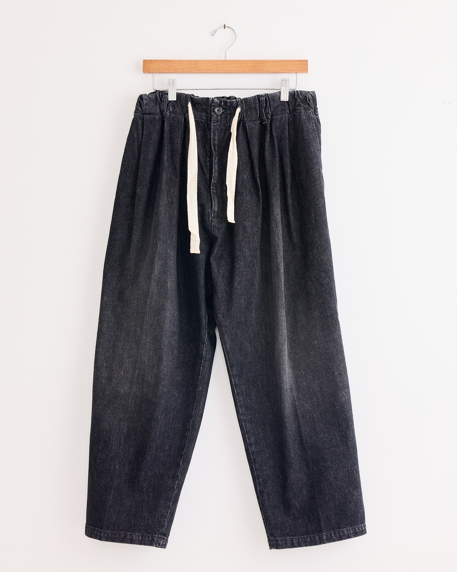 The Sillage Baggy Trouser Black in Denim Damaged, featuring an elastic waistband and white drawstring, hangs on a wooden hanger against a plain white wall.
