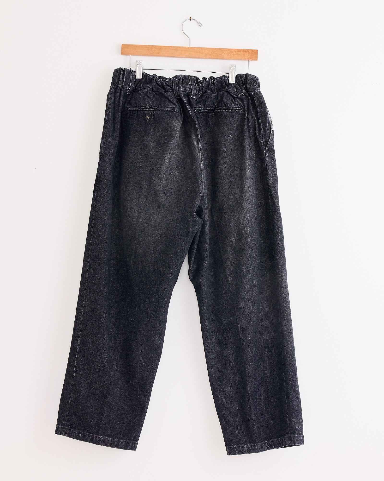 A pair of Sillage Baggy Trouser Black in Denim Damaged with an elastic waistband hangs on a wooden hanger against a plain white wall.