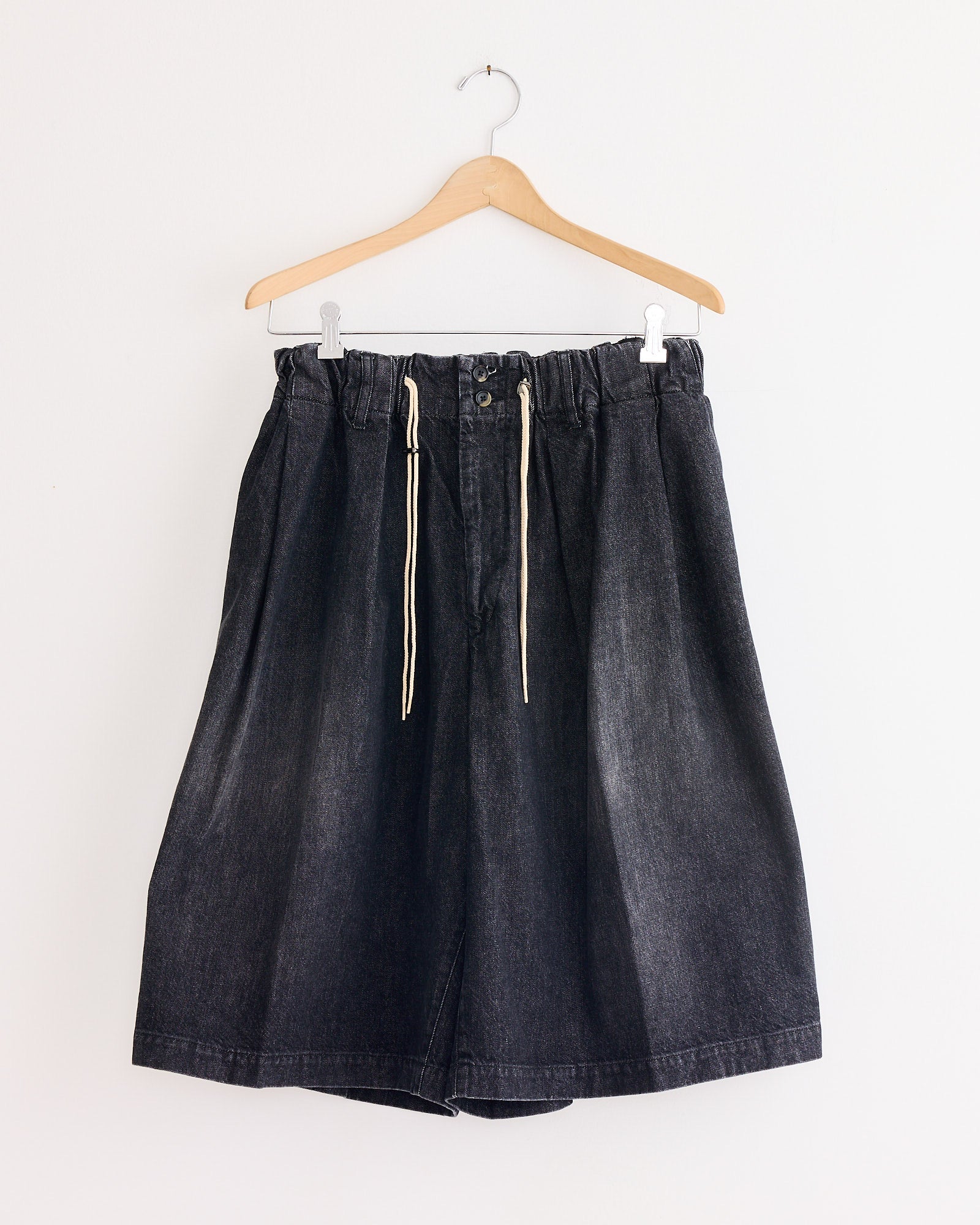 A pair of Sillage Circular Short Pant Black in Denim Damaged, featuring an elastic waistband, white drawstrings, and button closure, hangs on a wooden hanger against a white wall.