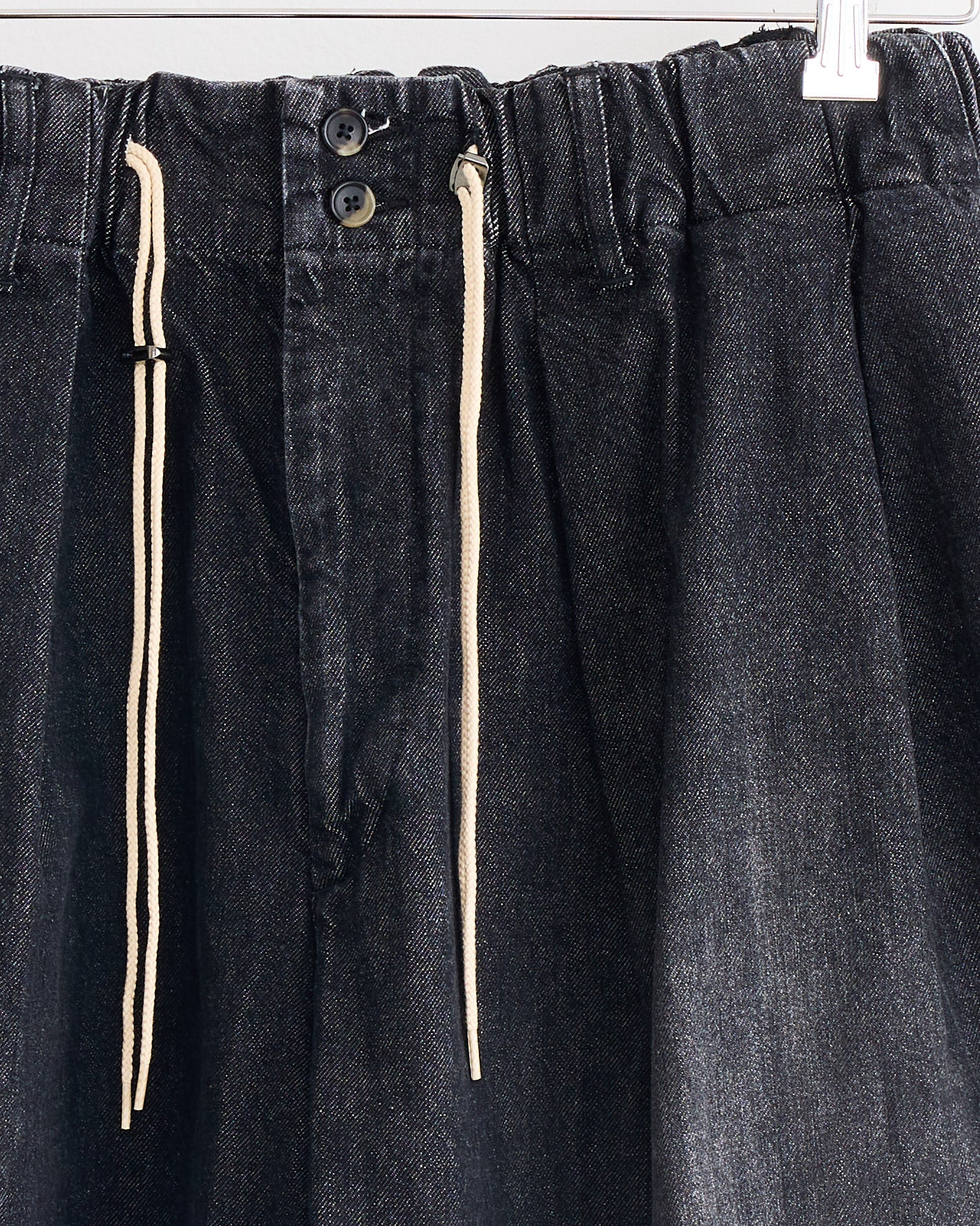 Close-up of Sillage Circular Short Pant Black in Denim Damaged, made from organic cotton with an elastic waistband, two black buttons, and white drawstrings. Made in Japan and shown on a hanger.
