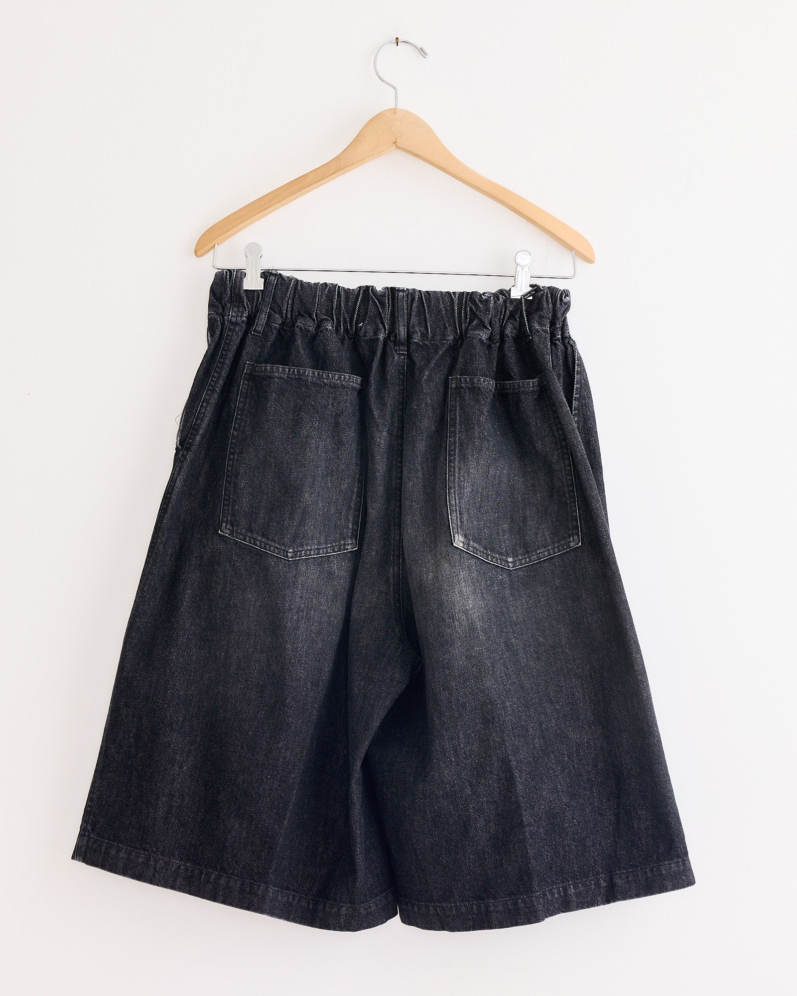 The Sillage Circular Short Pant Black in Denim Damaged features a wide-leg design, organic cotton denim, elastic waistband, two back pockets, and slight fading, displayed on a wooden hanger against a white wall.