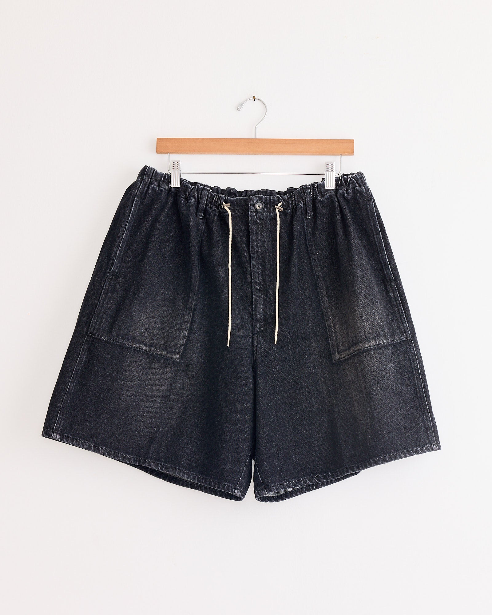 The Sillage Circular Fatigue Short Black in Denim Damaged features large front pockets, a white drawstring waistband, and is crafted from organic cotton denim in Japan. Displayed on a wooden hanger against a plain white wall.