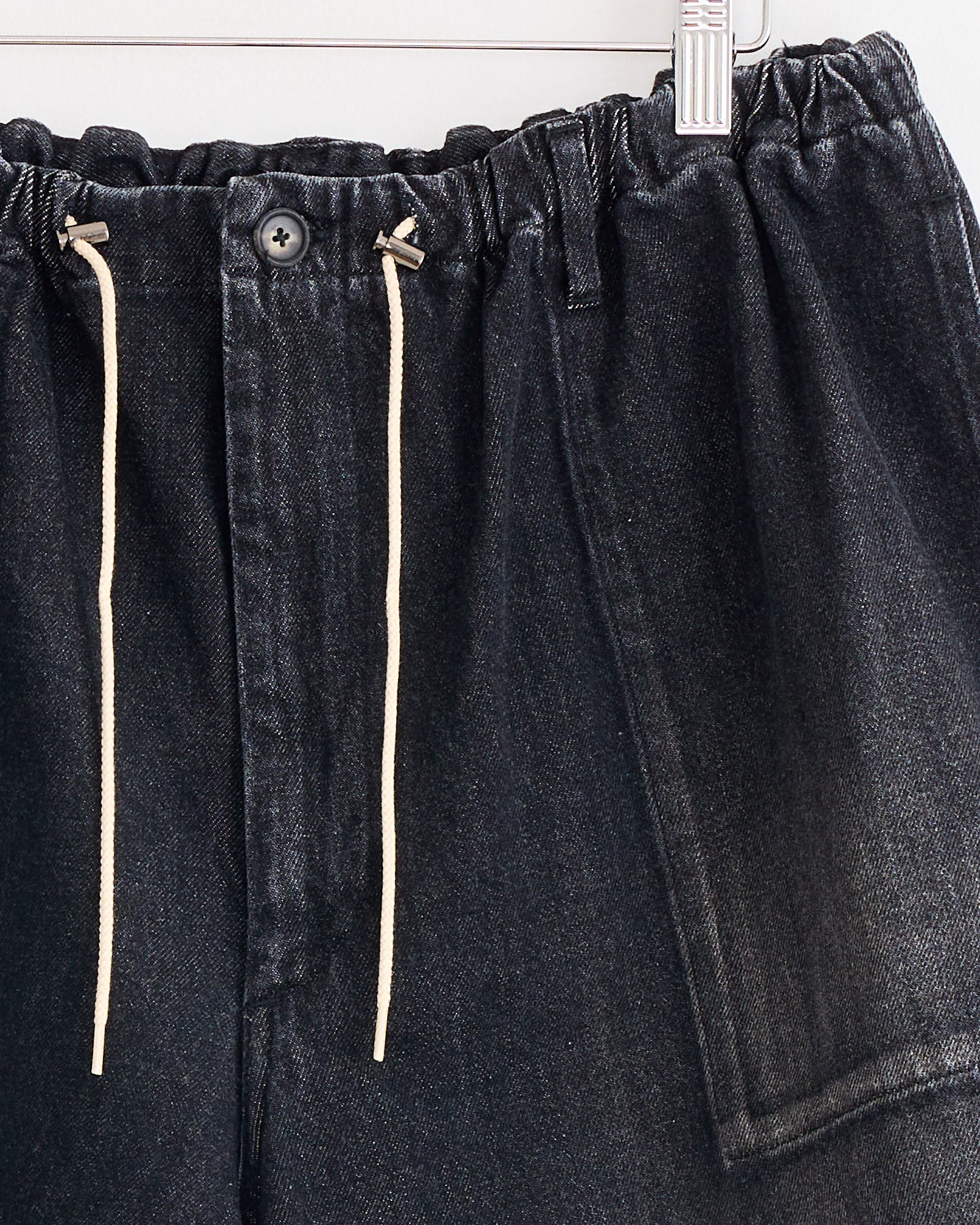 Close-up of Sillage Circular Fatigue Short Black in Denim Damaged—dark organic cotton denim shorts with elastic waistband, beige drawstrings, a button closure, and a large right pocket, made in Japan and hanging on a hanger.
