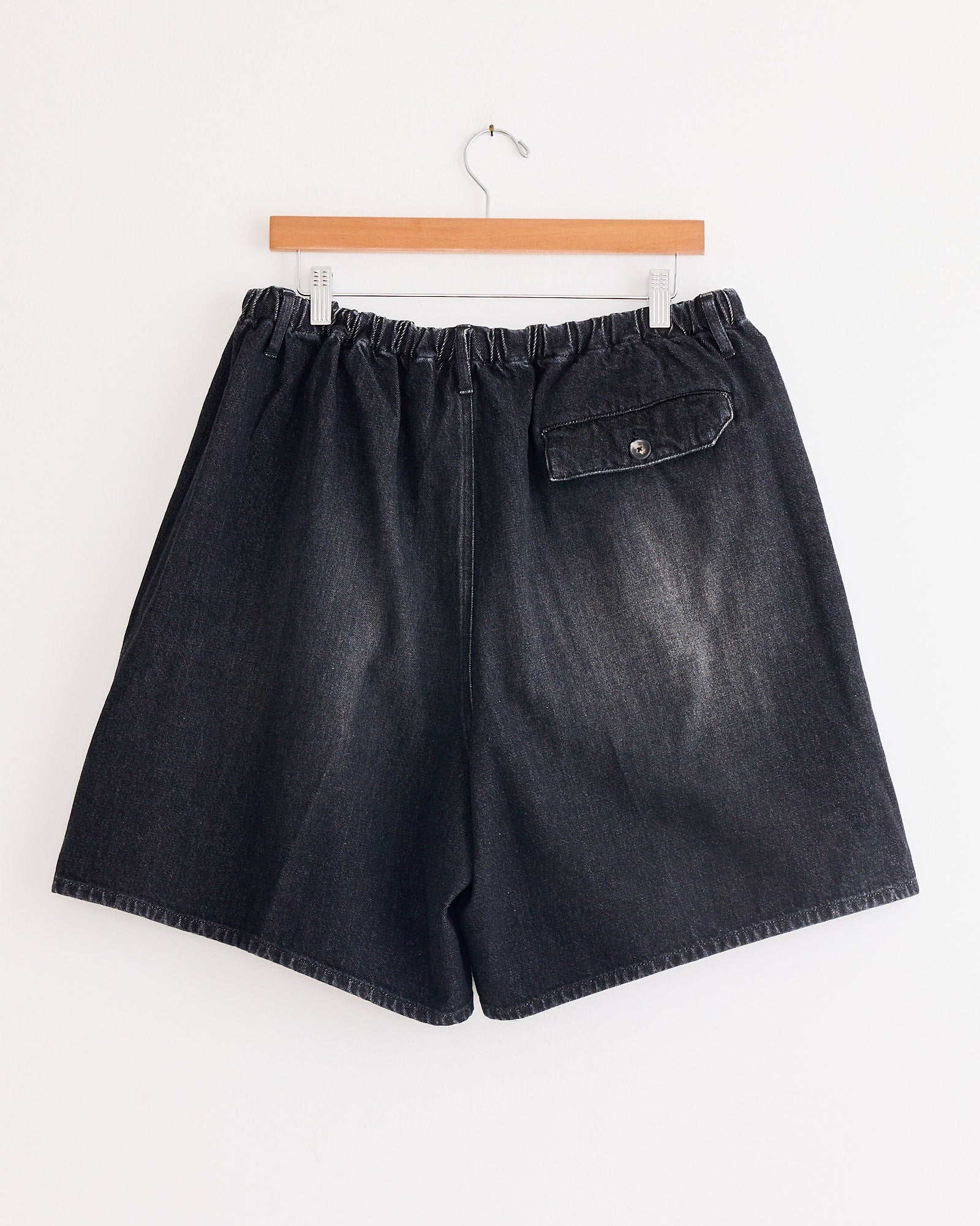The Sillage Circular Fatigue Short Black in Denim Damaged, made in Japan, features a loose fit, elastic waistband, and a single buttoned flap back pocket, shown hanging on a wooden hanger against a white wall.
