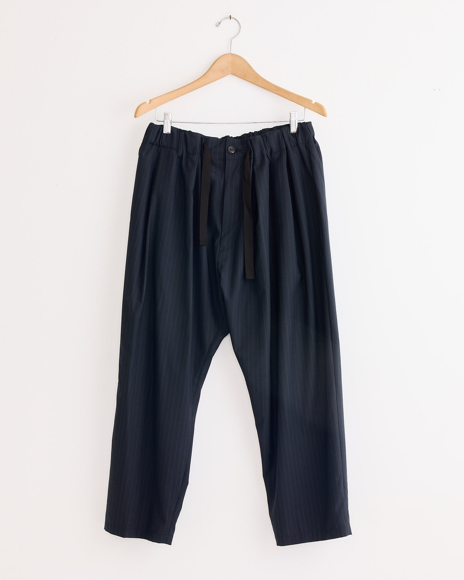 The Sillage Baggy Tropical Wool Trouser, featuring a drawstring and button waist, is displayed on a wooden hanger against a white wall. Made in Japan.