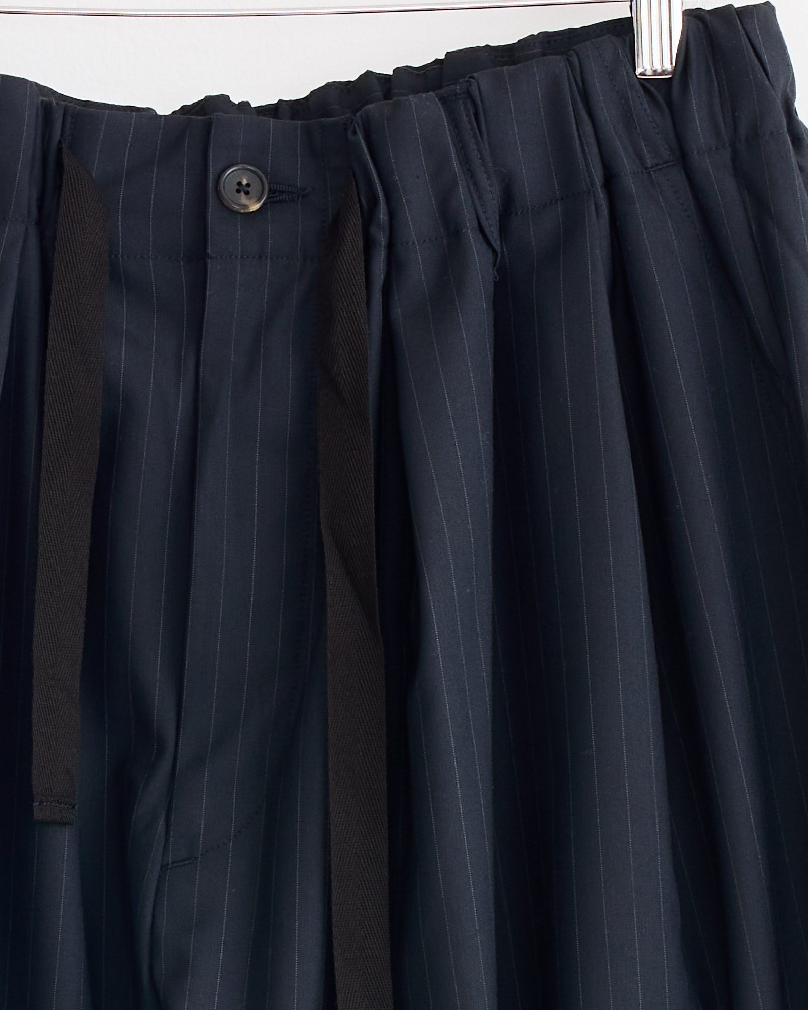 Close-up of Sillage Baggy Tropical Wool Trouser in navy pinstripe, featuring elastic waistband, black drawstring, and button. Crafted from tropical wool and hanging on a hanger. Made in Japan.