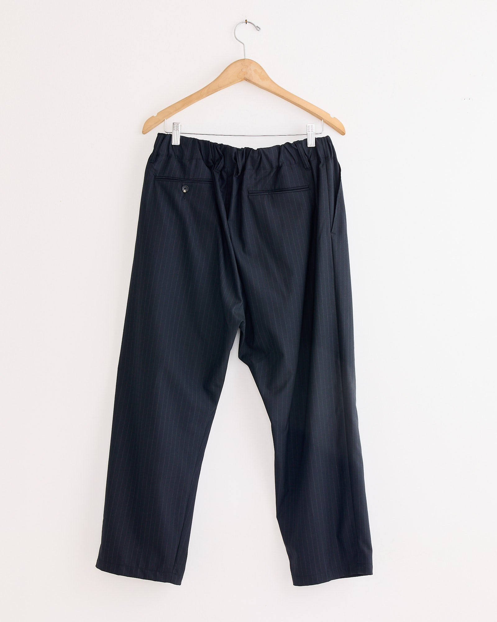 The Sillage Baggy Tropical Wool Trouser in black, made in Japan, hangs on a wooden hanger against a white wall. Featuring an elastic waistband, two back pockets, and a button closure on one pocket.