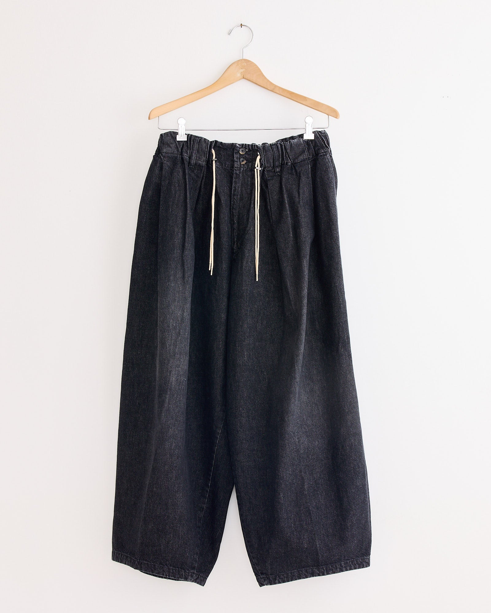 The Sillage Circular Pant Black in Denim Damaged, with a wide-leg silhouette, elastic waistband, and white drawstrings, hangs on a wooden hanger against a plain white wall—blending modern comfort with iconic style.
