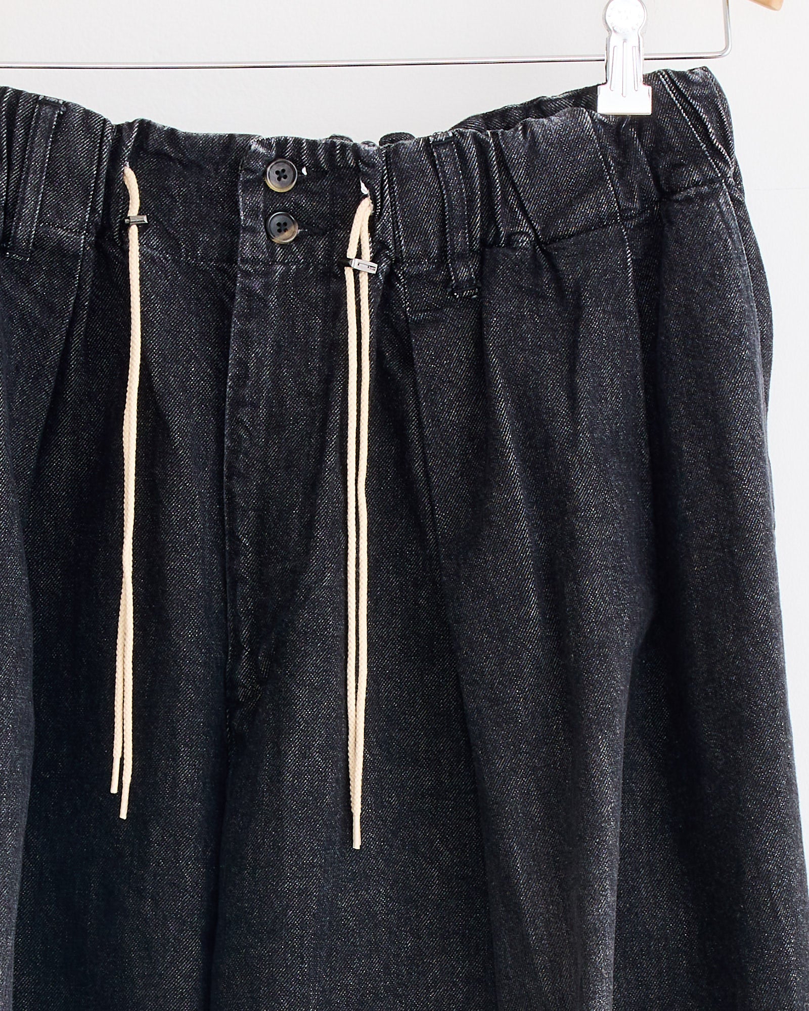 A close-up of Sillage's Circular Pant Black in Denim Damaged, featuring an elastic waistband, two black buttons, and off-white drawstrings, hanging on a hanger against a light background.