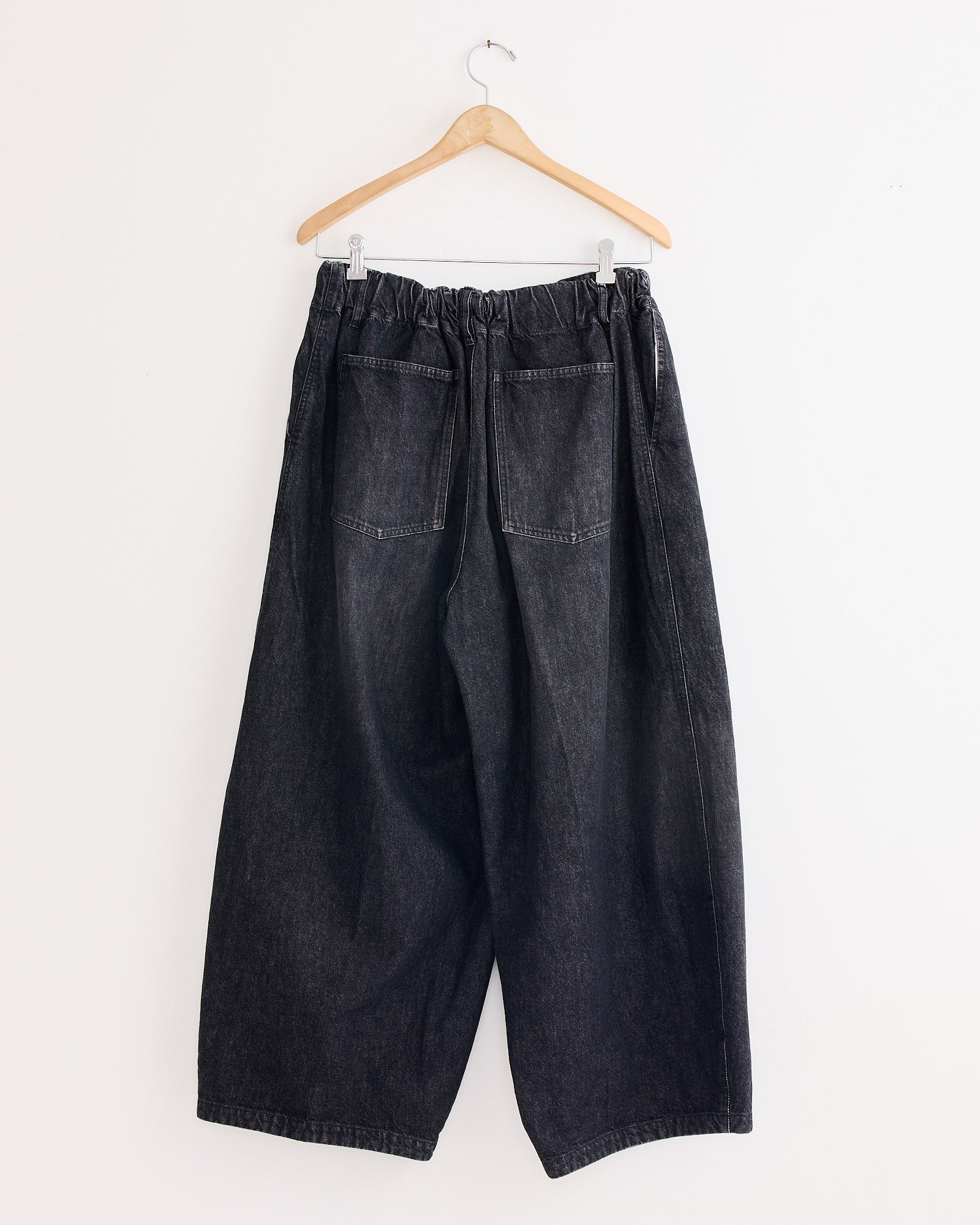 The Sillage Circular Pant Black in Denim Damaged features a loose fit, elastic waistband, and two back pockets, displayed on a wooden hanger against a plain white wall.