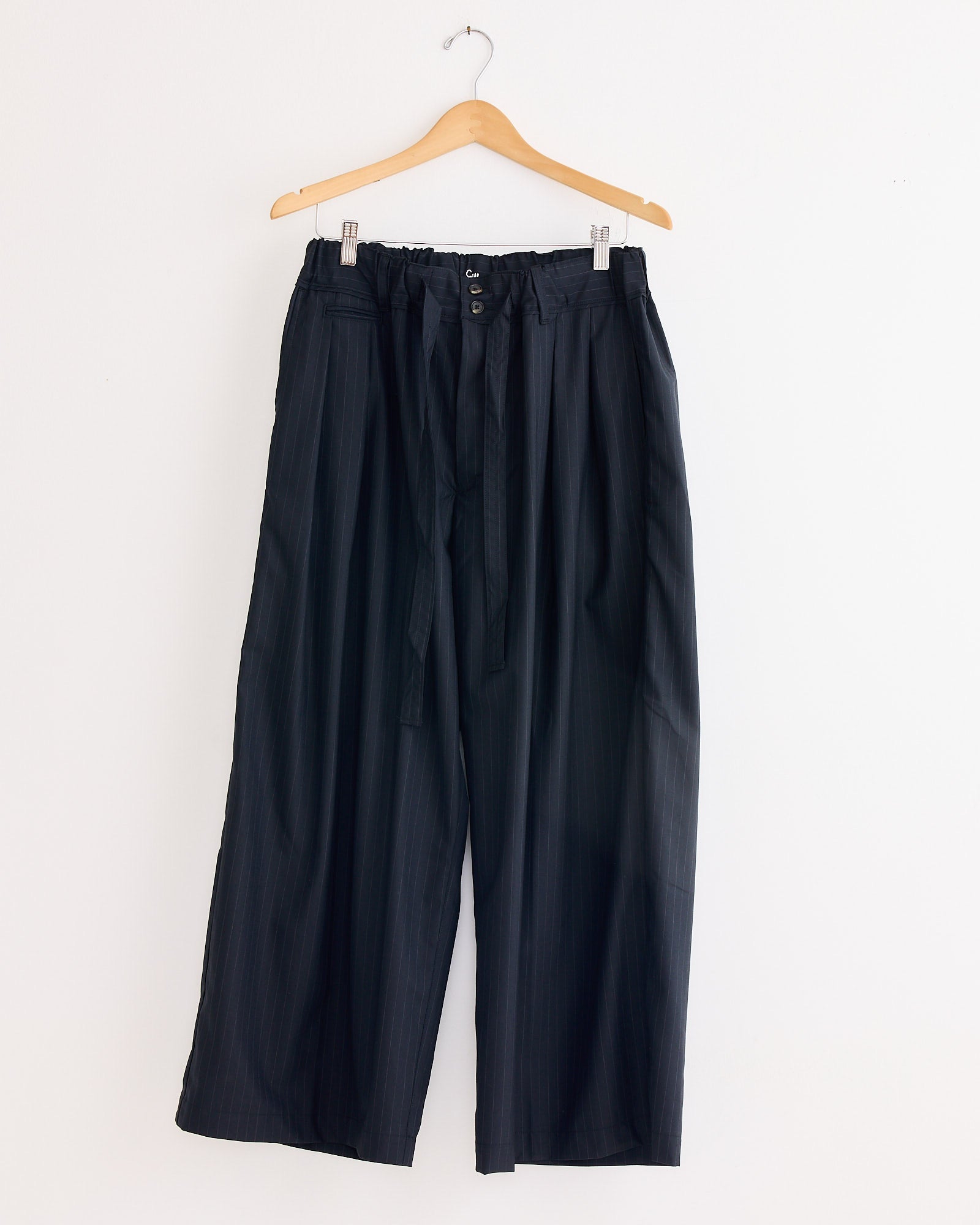 A pair of Sillage Hakama Tropical Wool Pants hangs on a wooden hanger against a white wall, featuring a high waist, wide silhouette, paperbag waist design, drawstring tie, and button closure.