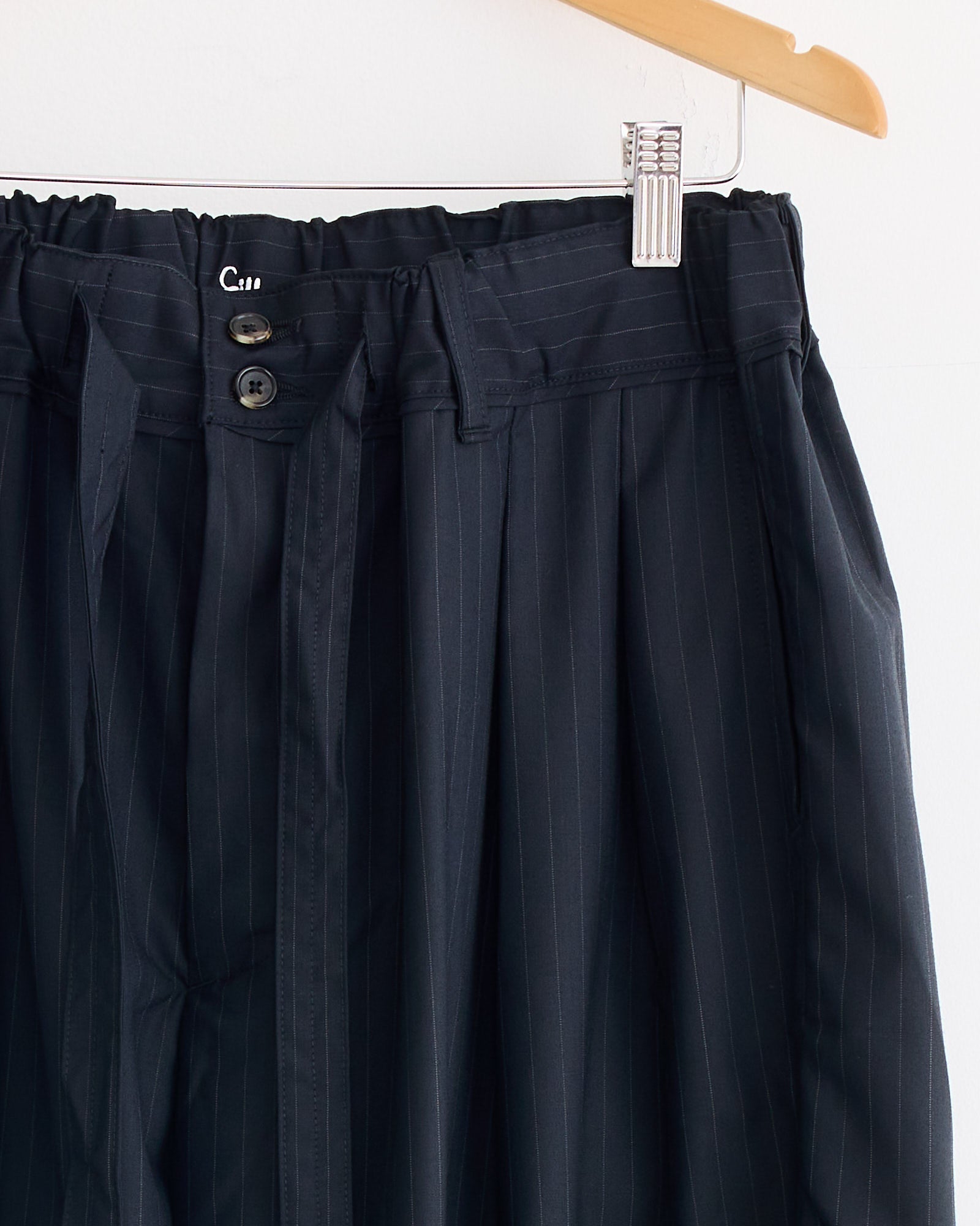 The Sillage Hakama Tropical Wool Pant hangs on a wooden hanger, featuring a wide straight silhouette, paperbag waist with belt loops, fabric belt, and two black button closures.