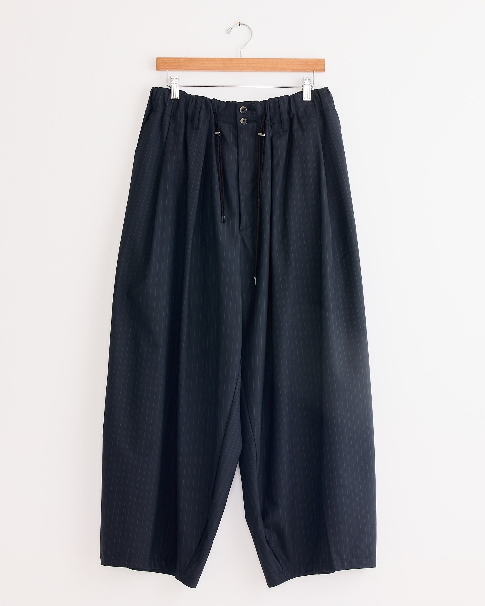 The Sillage Circular Tropical Wool Pant in Black Stripe, made in Japan, features a loose fit, elastic waistband, and drawstring. Shown hanging on a wooden hanger against a white wall.