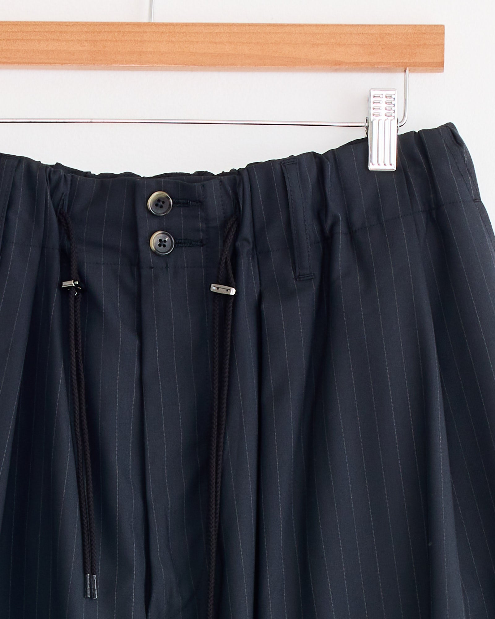 Close-up of the waistband of Sillage Circular Tropical Wool Pant in Black Stripe, made in Japan. Shown on a wooden hanger with two buttons, an elastic waistband, and black drawstrings with metal tips.