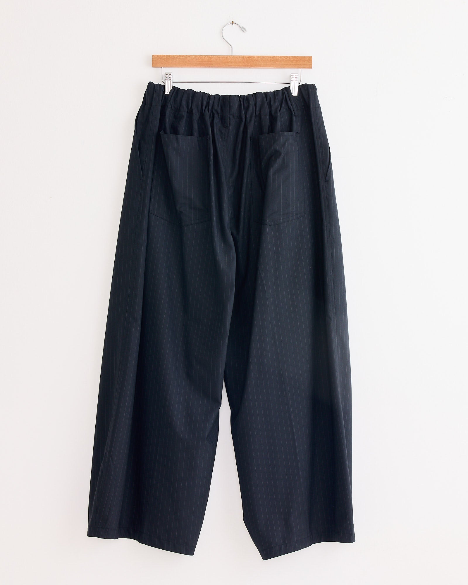 The Sillage Circular Tropical Wool Pant in Black Stripe, featuring a loose wide-leg fit, elastic waistband, and two large back pockets, is made in Japan and hangs on a wooden hanger against a white wall.