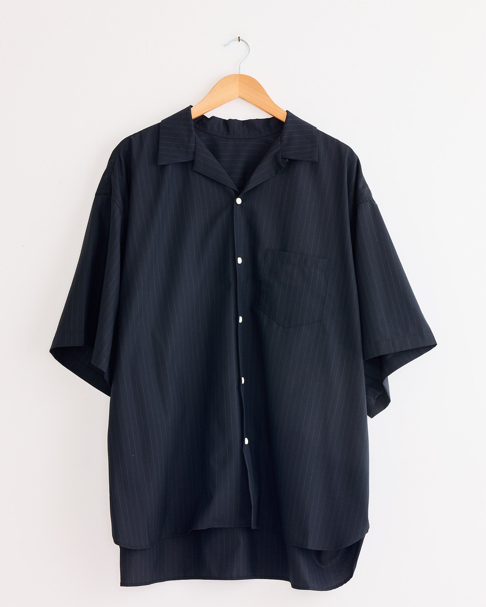 The Sillage Re-Engineered Tropical Wool S/S Overshirt, featuring subtle pinstripes, a collar, and chest pocket, hangs on a wooden hanger against a white wall.