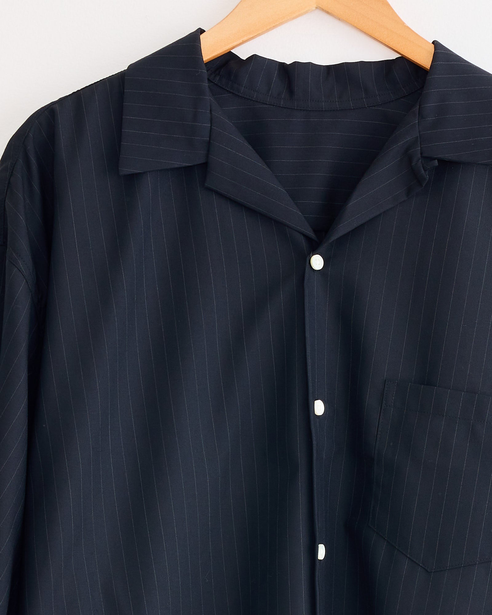 A close-up of the Sillage Re-Engineered Tropical Wool S/S Overshirt in dark pinstripe on a wooden hanger, with white buttons, a front pocket, and a pointed collar against a light background.