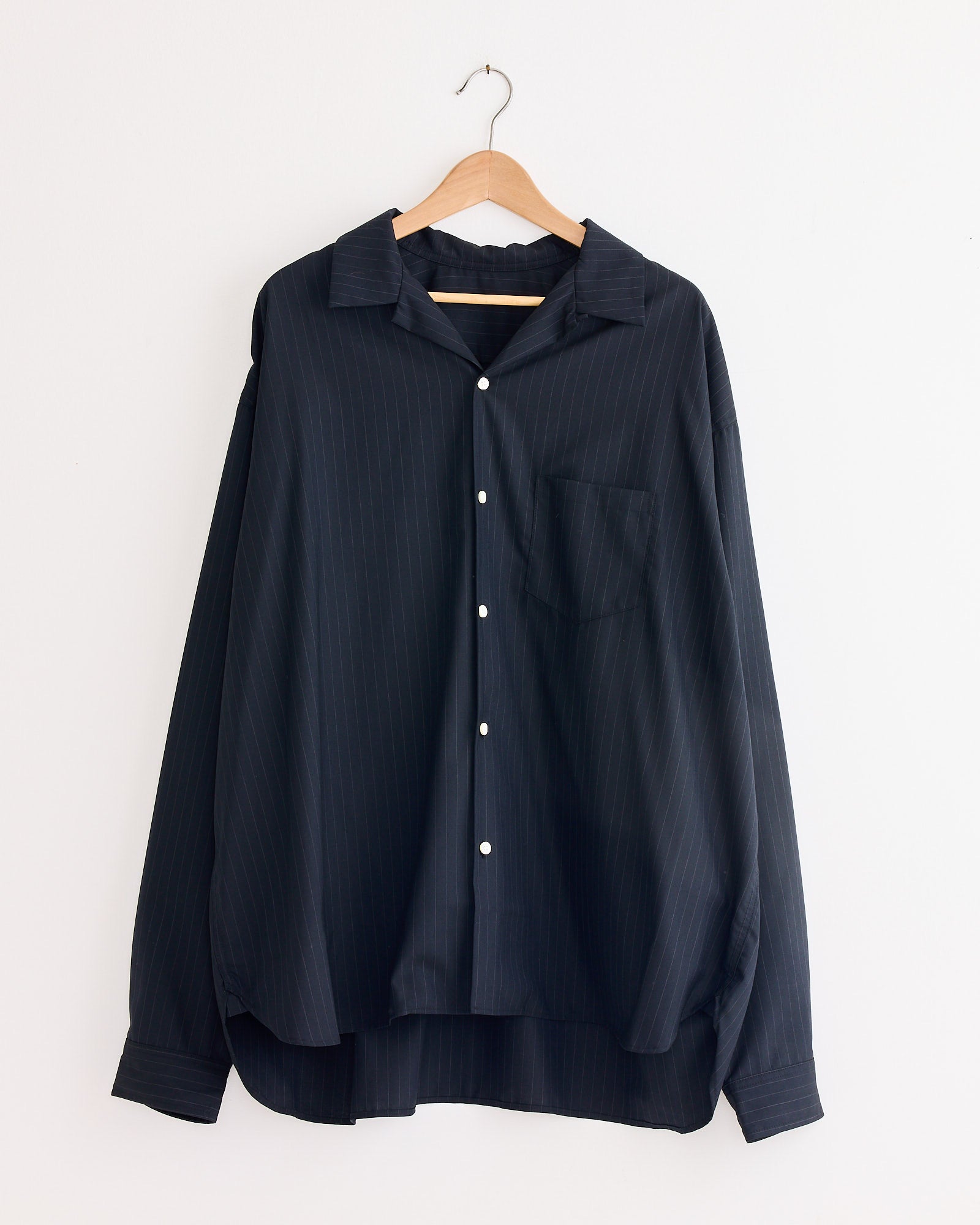 The Sillage Re-Engineered Tropical Wool L/S Overshirt in Stripe, a loose-fitting dark navy piece with subtle vertical pinstripes, hangs on a wooden hanger against a plain white wall.