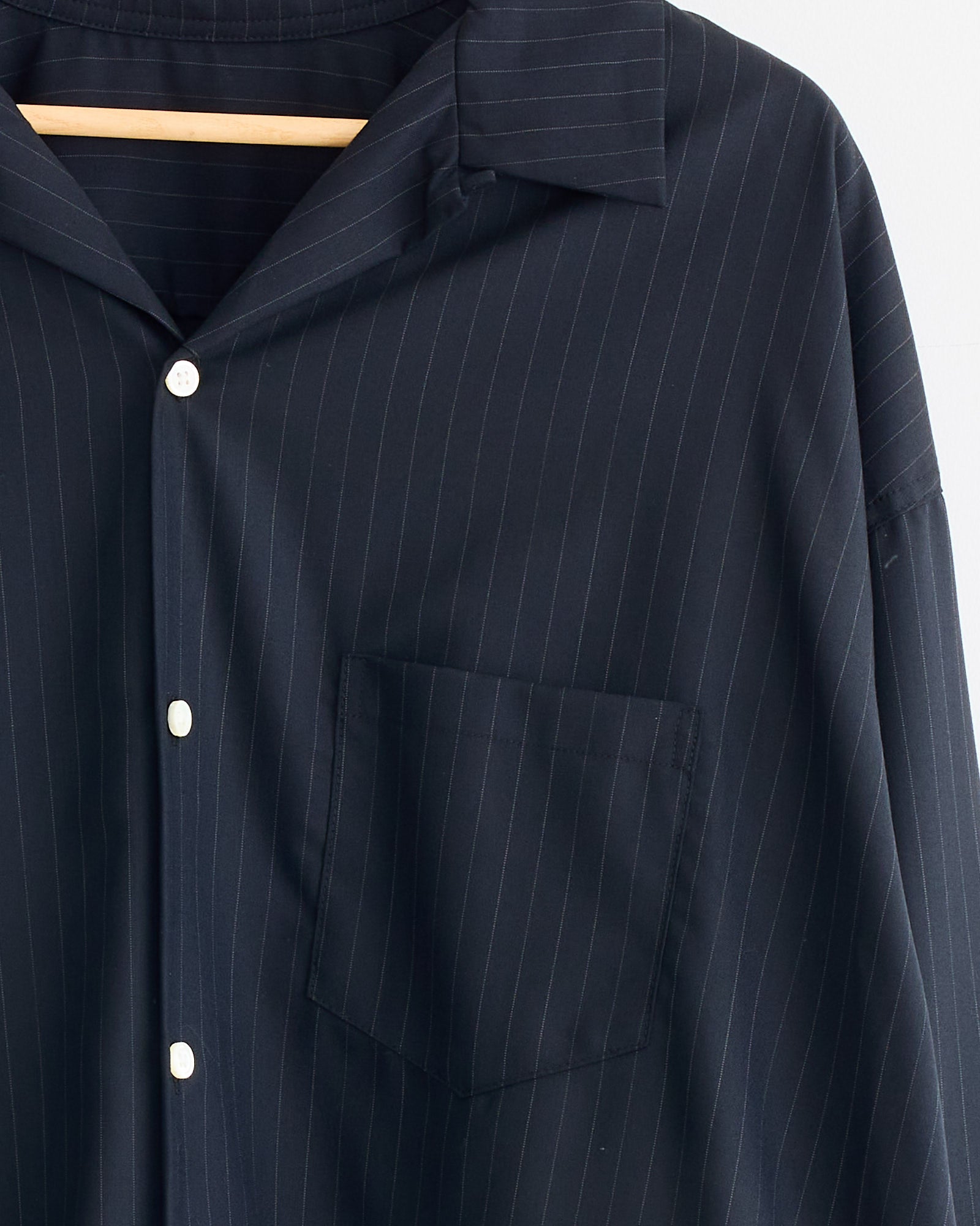 A close-up of the Sillage Re-Engineered Tropical Wool L/S Overshirt in Stripe, featuring dark navy pinstripes, white buttons, a pointed collar, and chest pocket, displayed on a wooden hanger.