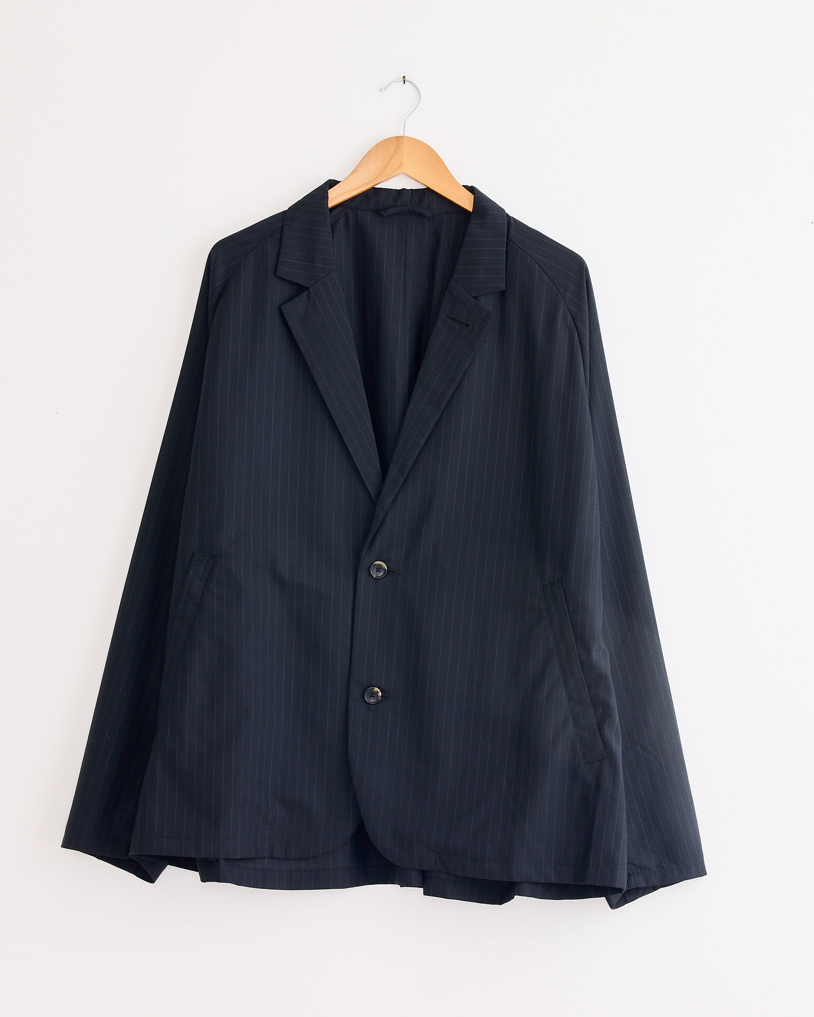 The Sillage Veste Tropical Wool Two Button Jacket in Black Stripe hangs on a wooden hanger against a plain white wall.