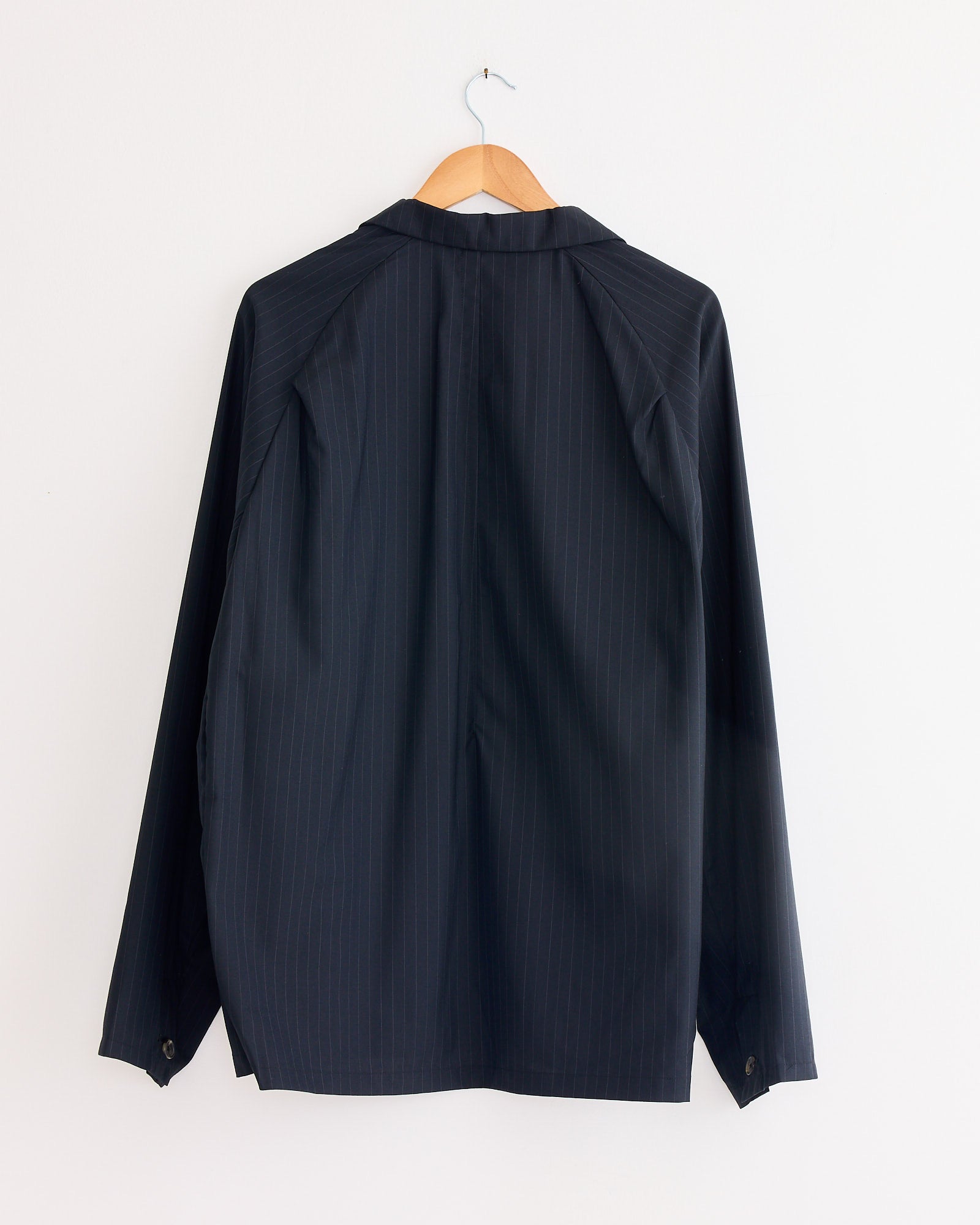 The Sillage Veste Tropical Wool Two Button Jacket in Black Stripe, shown from the back, features dark pinstripes and long sleeves as it hangs on a wooden hanger against a plain white wall.