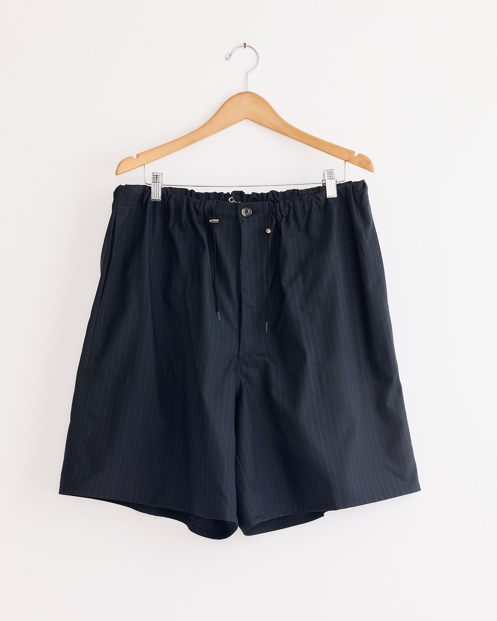 The Sillage Circular Fatigue Short Pant in black pinstriped tropical wool features an elastic waistband, drawstring ties, and button closure. Shown on a wooden hanger against a white wall. Made in Japan.