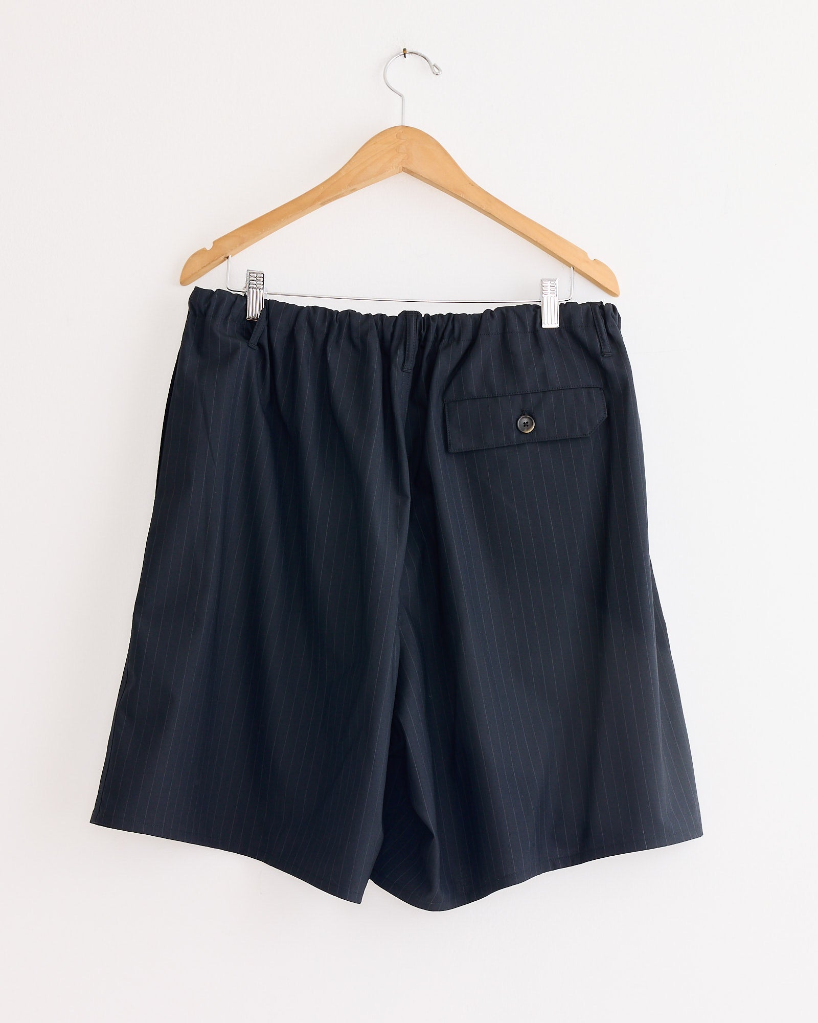 A pair of Sillage Circular Fatigue Short Pant in black pinstripe tropical wool with an elastic waistband and buttoned back pocket hangs on a wooden hanger against a white wall. Made in Japan.
