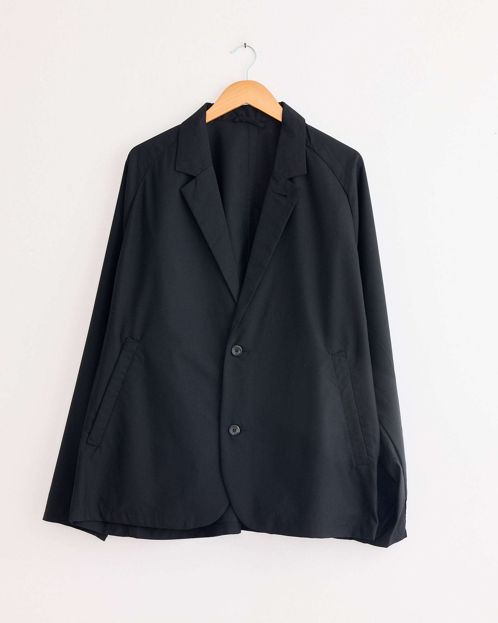 The Sillage Veste Tropical Wool Two Button Jacket in Black hangs on a wooden hanger against a plain white wall.