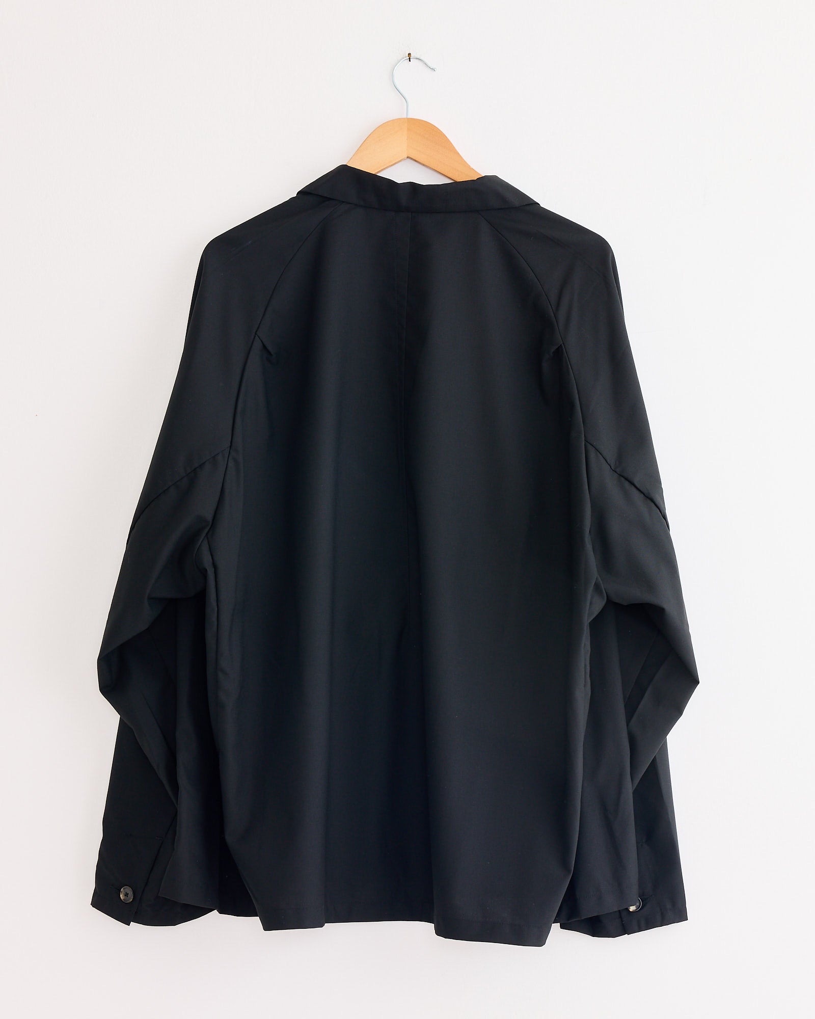 The Sillage Veste Tropical Wool Two Button Jacket in Black hangs on a wooden hanger against a plain white wall, shown from the back.