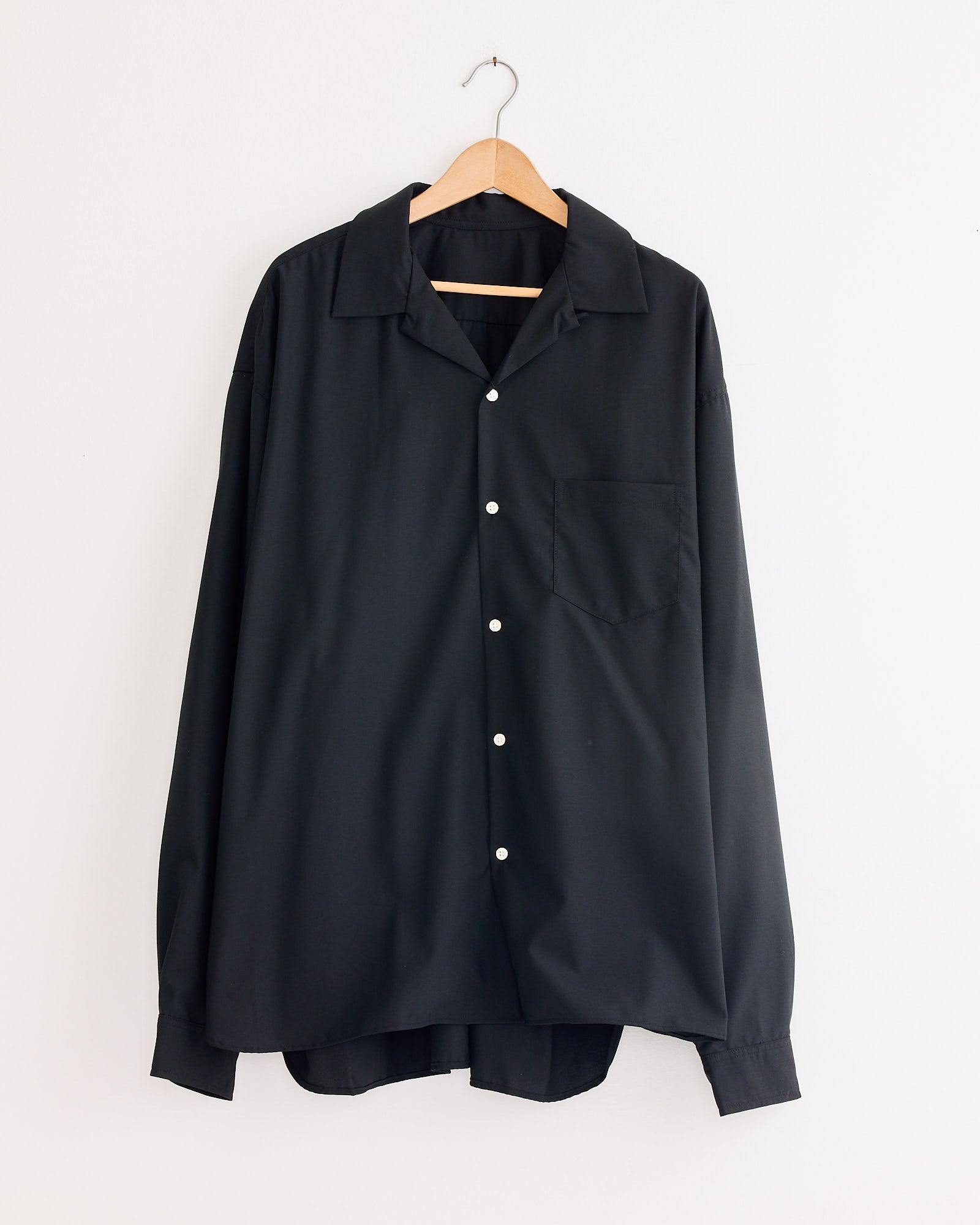 The Sillage Re-Engineered Tropical Wool L/S Overshirt in Black, featuring a collar and chest pocket, hangs on a wooden hanger against a white wall.