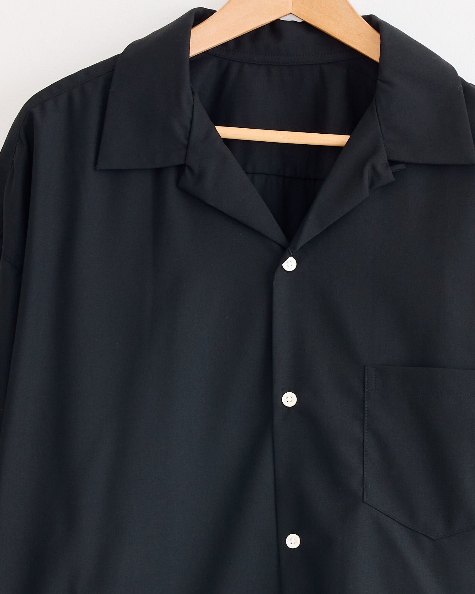 The Sillage Re-Engineered Tropical Wool L/S Overshirt in Black, featuring a collar and chest pocket, hangs on a wooden hanger against a plain white background—ideal as a lightweight layer for any occasion.
