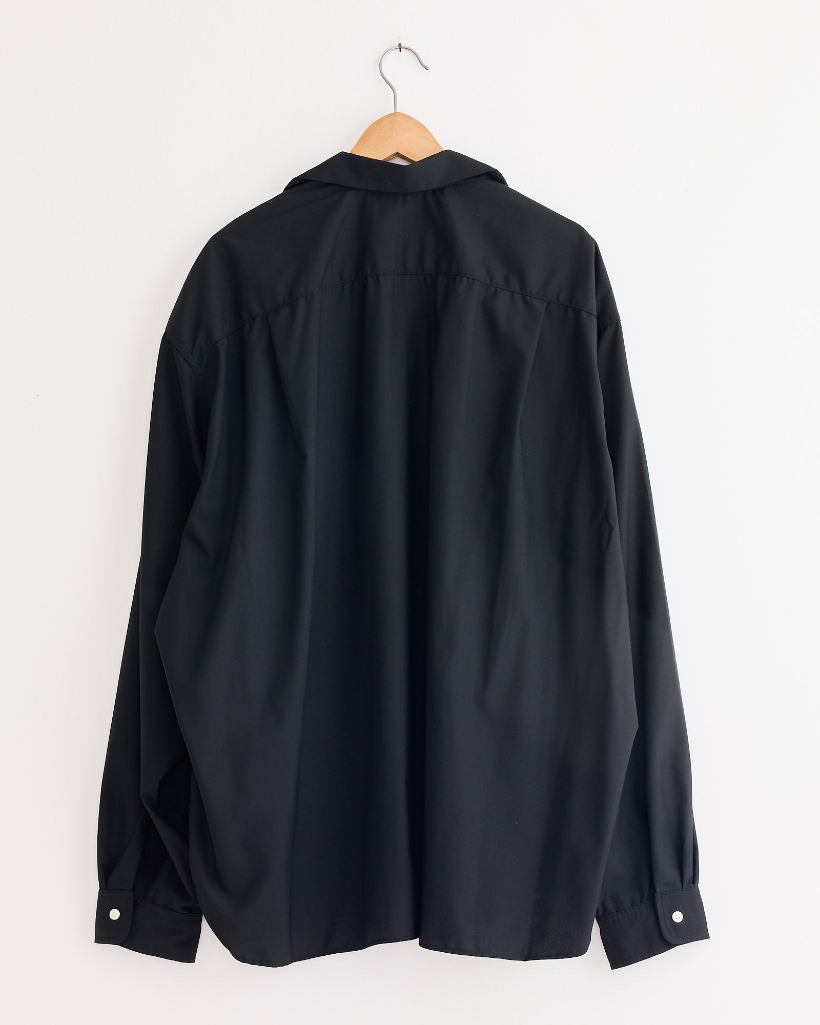 A Re-Engineered Tropical Wool L/S Overshirt in Black by Sillage hangs on a wooden hanger against a plain white wall, displayed from the back with loosely hanging sleeves and buttoned cuffs.