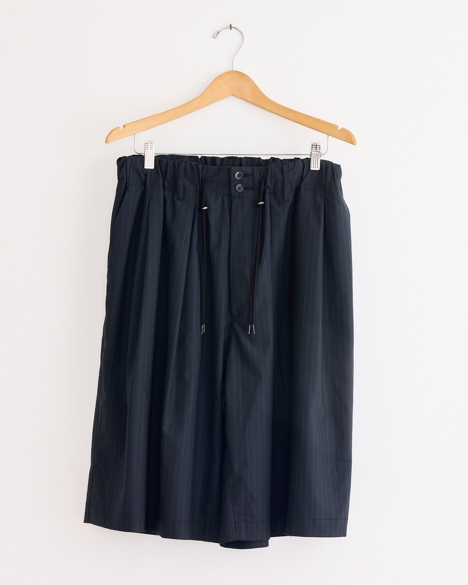 The Sillage Circular Short Tropical Wool Pant in Black Stripe features a wide circular cut, knee length, elastic waistband with drawstring and buttons, shown hanging on a wooden hanger against a white wall.