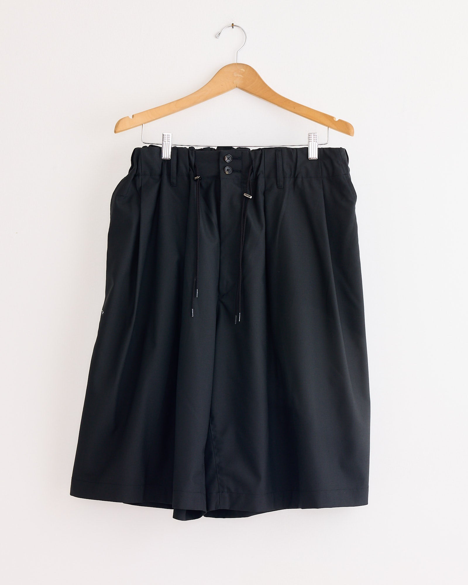 A pair of Sillage Circular Short Tropical Wool Pants in Black, featuring a loose fit, wide circular cut, elastic waistband, and drawstring, hang on a wooden hanger against a plain white wall.