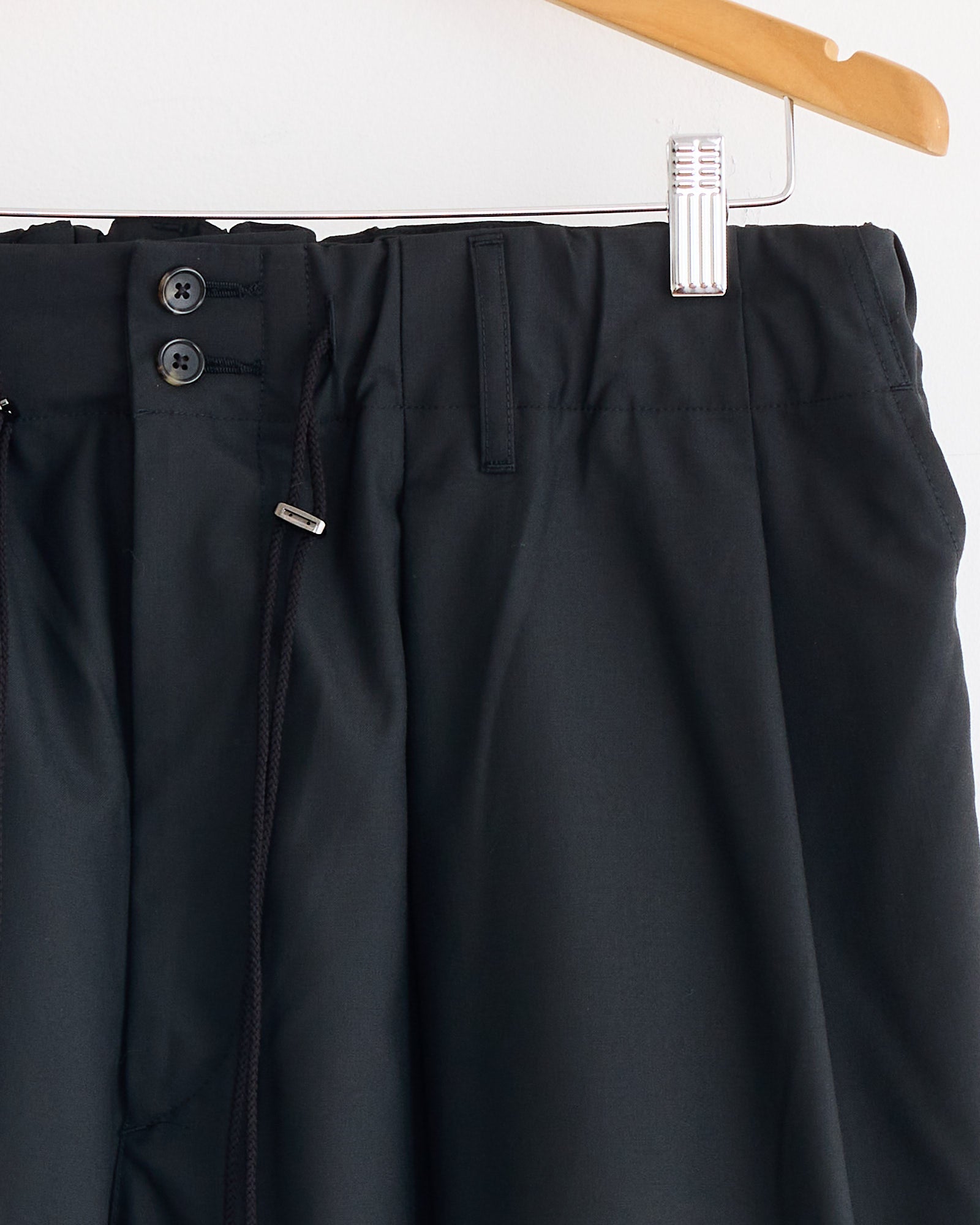 Close-up of the waistband of Sillage’s Circular Short Tropical Wool Pant in Black, showing two black buttons, belt loops, a partially visible zipper, elastic waistband with a black drawstring. Made in Japan and hung on a wooden hanger.