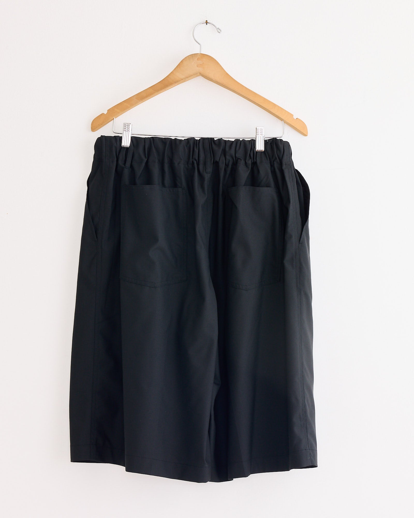 The Sillage Circular Short Tropical Wool Pant in Black, featuring a wide circular cut and large front pockets, is made in Japan and hangs on a wooden hanger against a plain white wall.