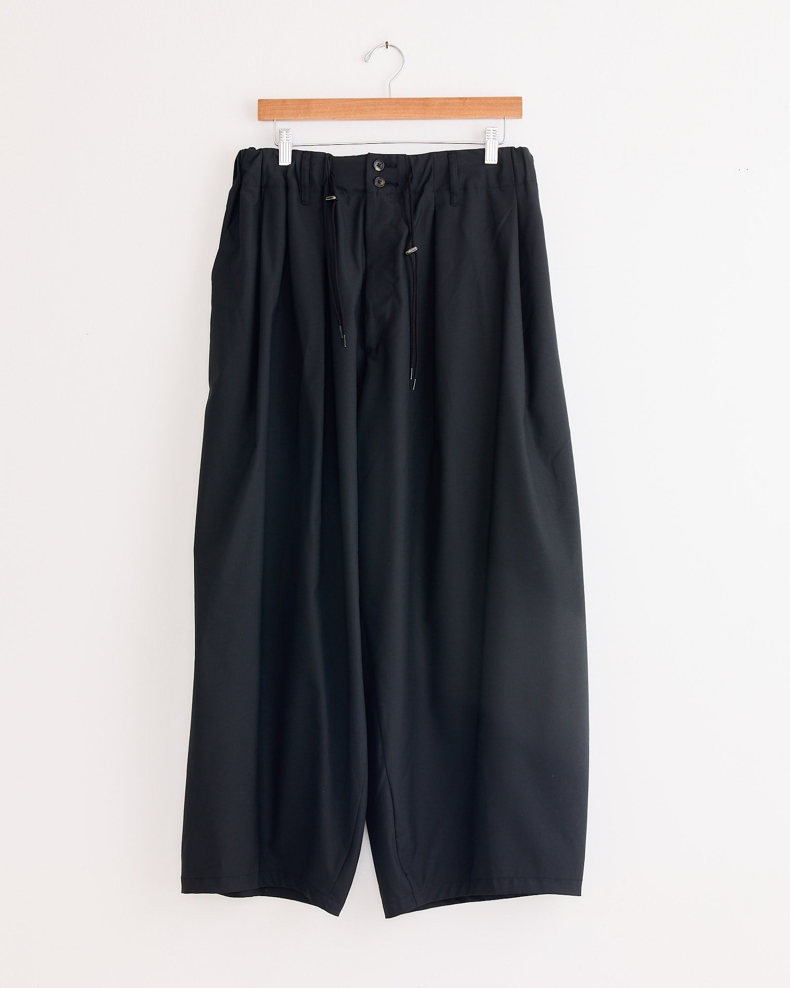 The Sillage Circular Tropical Wool Pant in Black, featuring a wide-leg silhouette, elastic waistband, drawstring, and belt loops, hangs on a wooden hanger against a white background.