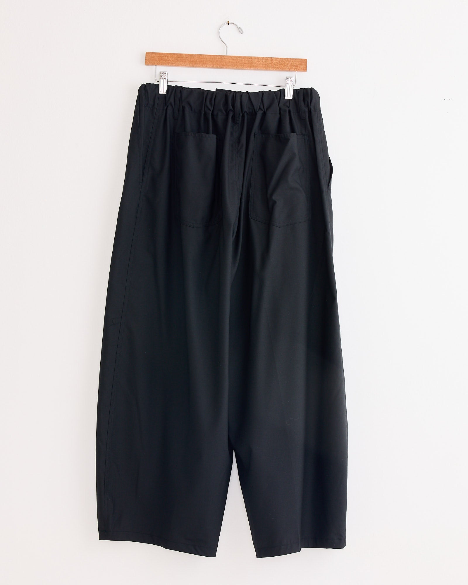 The Sillage Circular Tropical Wool Pant in Black, featuring an elastic waistband and back pockets, hangs on a wooden hanger against a white wall, highlighting its voluminous silhouette.
