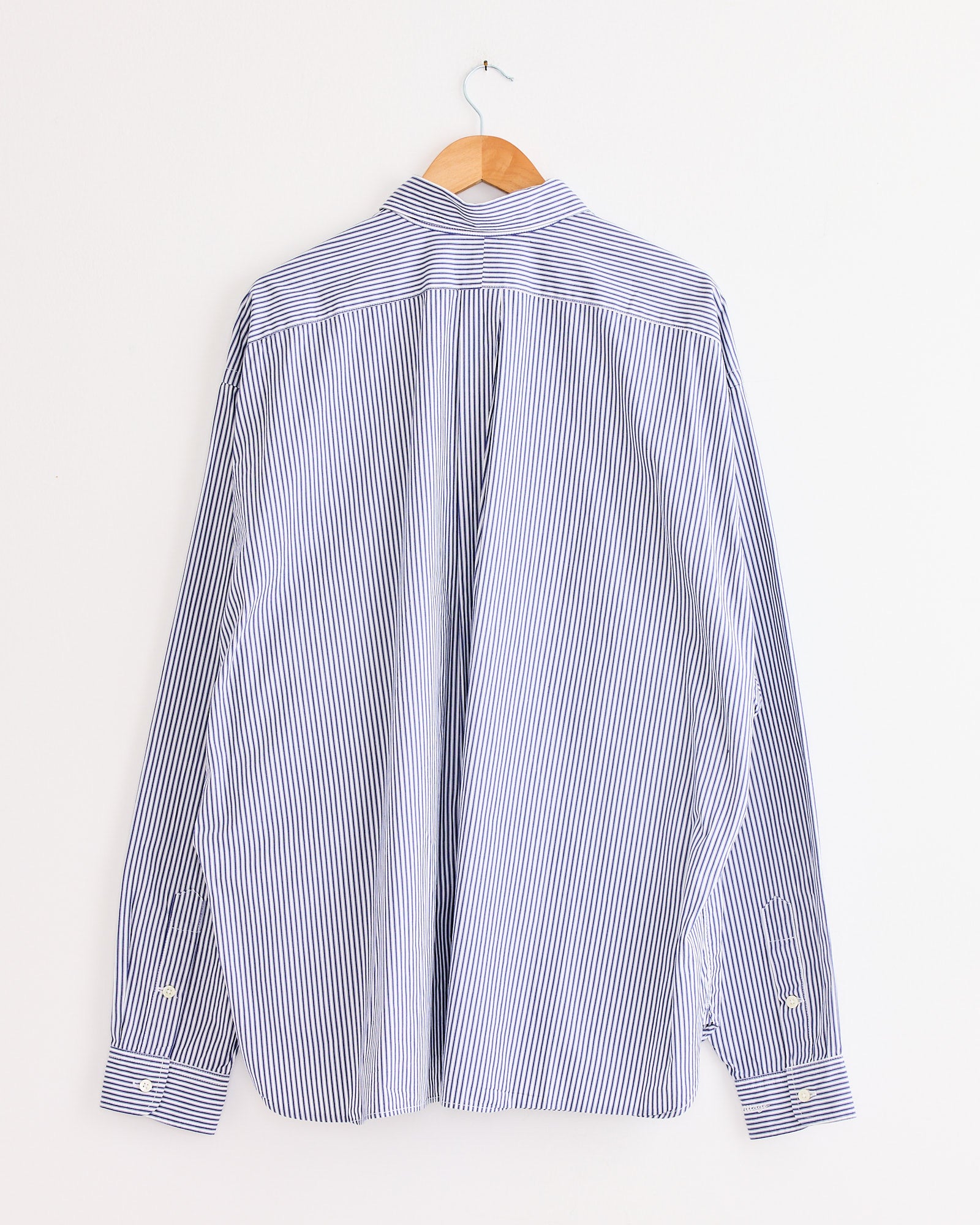 The Sillage Regular Wide High Count Shirt Navy Stripe, featuring blue and white vertical stripes and long sleeves, hangs on a wooden hanger against a plain white wall, shown from the back.