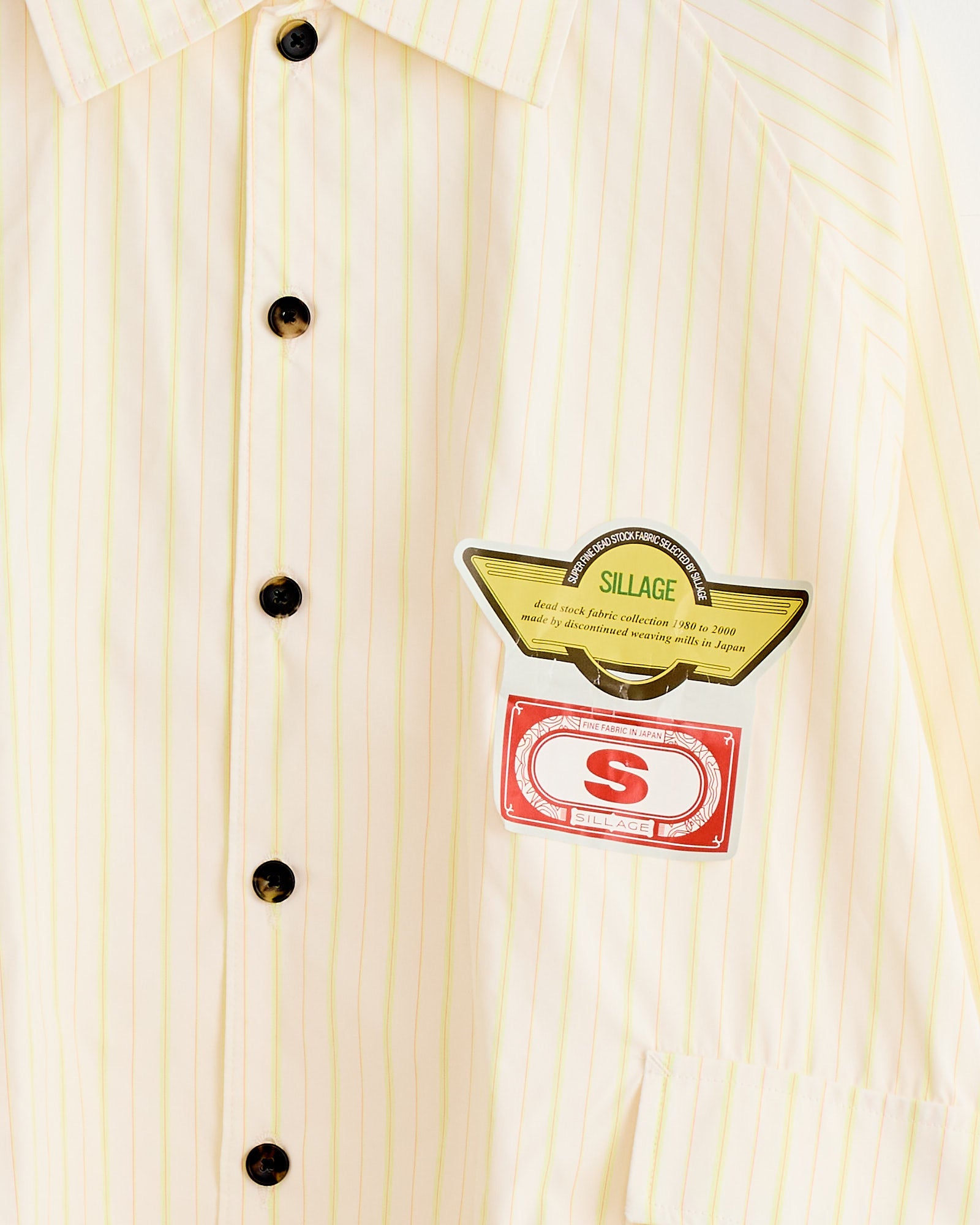 The Big Pocket Deadstock Overshirt by Sillage is cream-colored with pinstripes, black buttons, and two decorative patches: a yellow wing-shaped SILLAGE patch and a red S logo patch, both made from vintage deadstock materials.