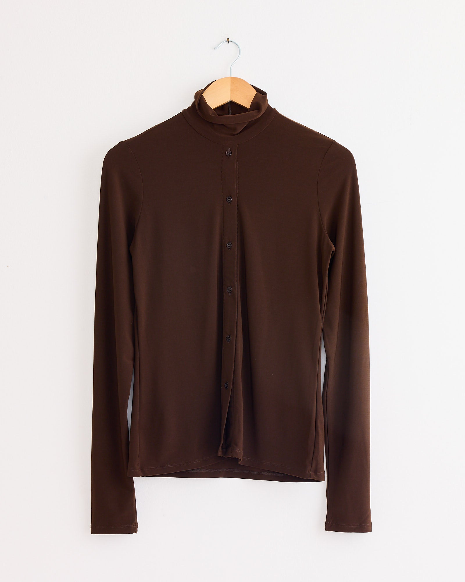 The Beaufille Button Down Rodin Blouse in Brown, featuring long sleeves, a high collar, and lightweight stretch fabric, hangs on a wooden hanger against a plain white wall.