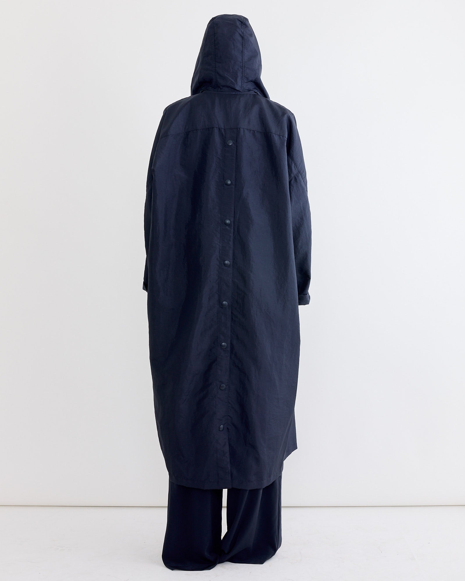 A person stands with their back turned, wearing the Tibi Crispy Nylon Hooded Coat in Navy—perfect transitional outerwear—over wide-legged pants, set against a plain white background.