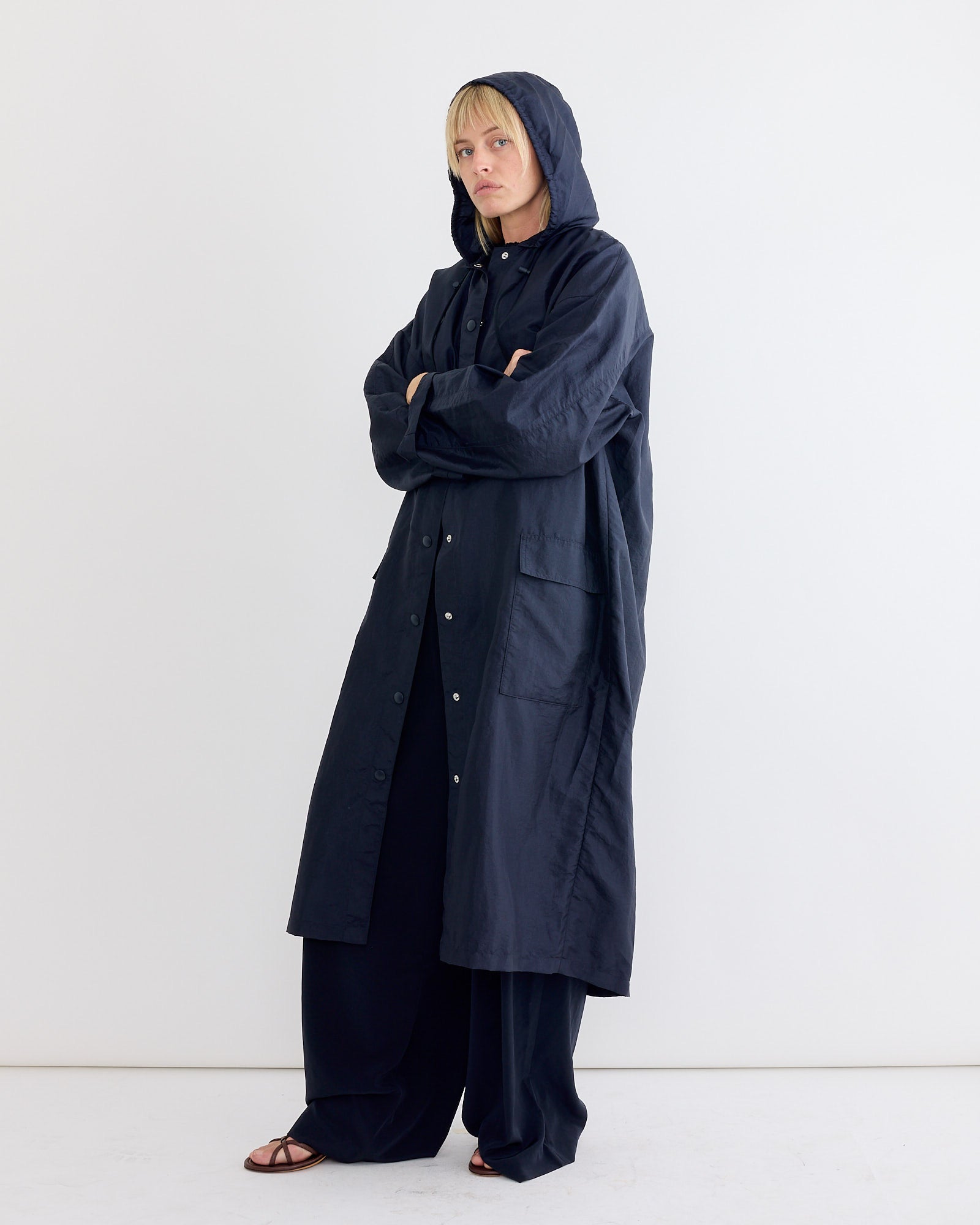A person stands against a plain white background, wearing the Tibi Crispy Nylon Hooded Coat in Navy over wide-leg pants and open-toe sandals. Their arms are crossed and their expression is neutral—perfect transitional outerwear.
