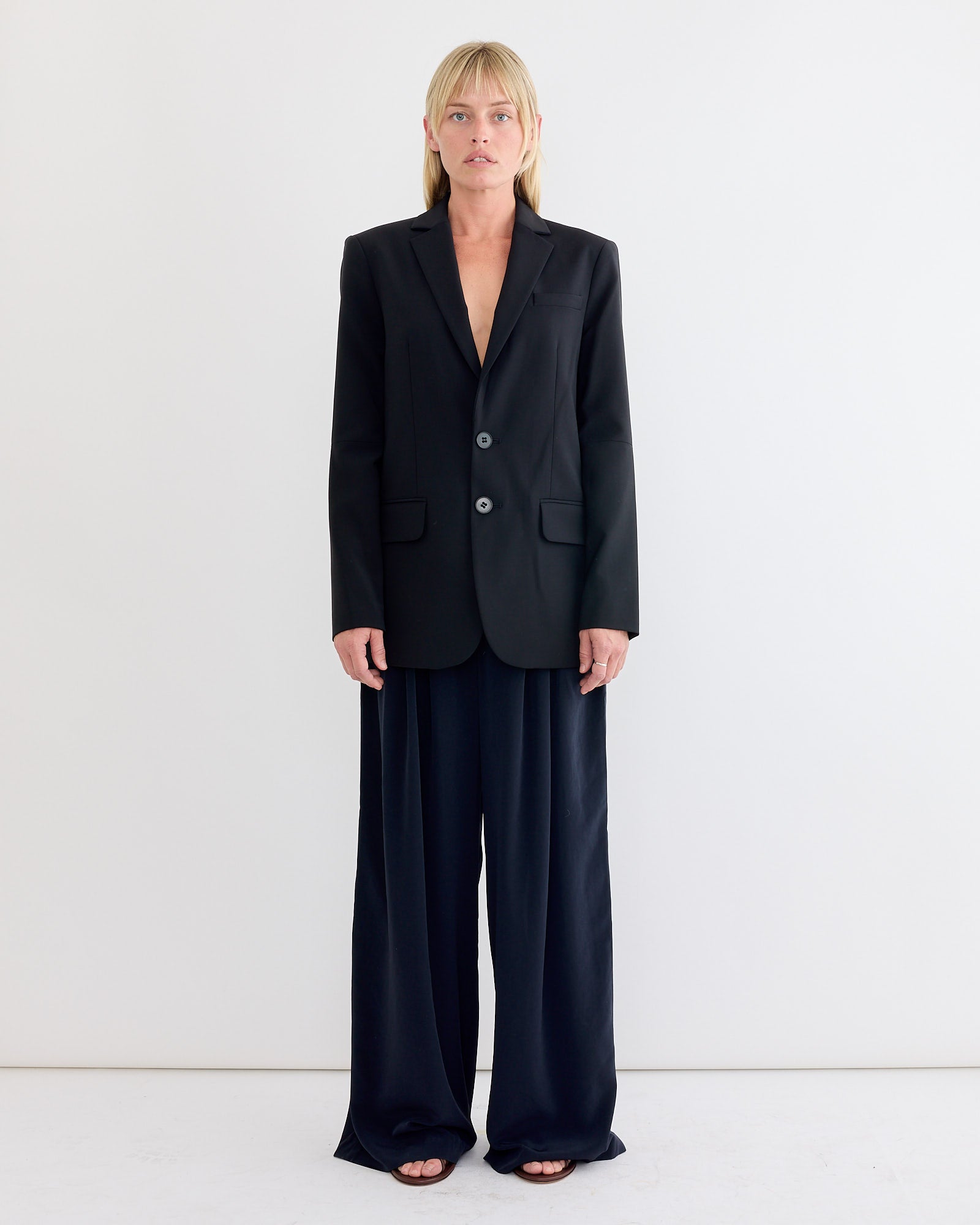 A person with straight blonde hair stands against a white background, wearing a black blazer, Tibi Relaxed Suiting Triple Pleat Pant in Navy, and open-toe black sandals for a minimal, modern look.