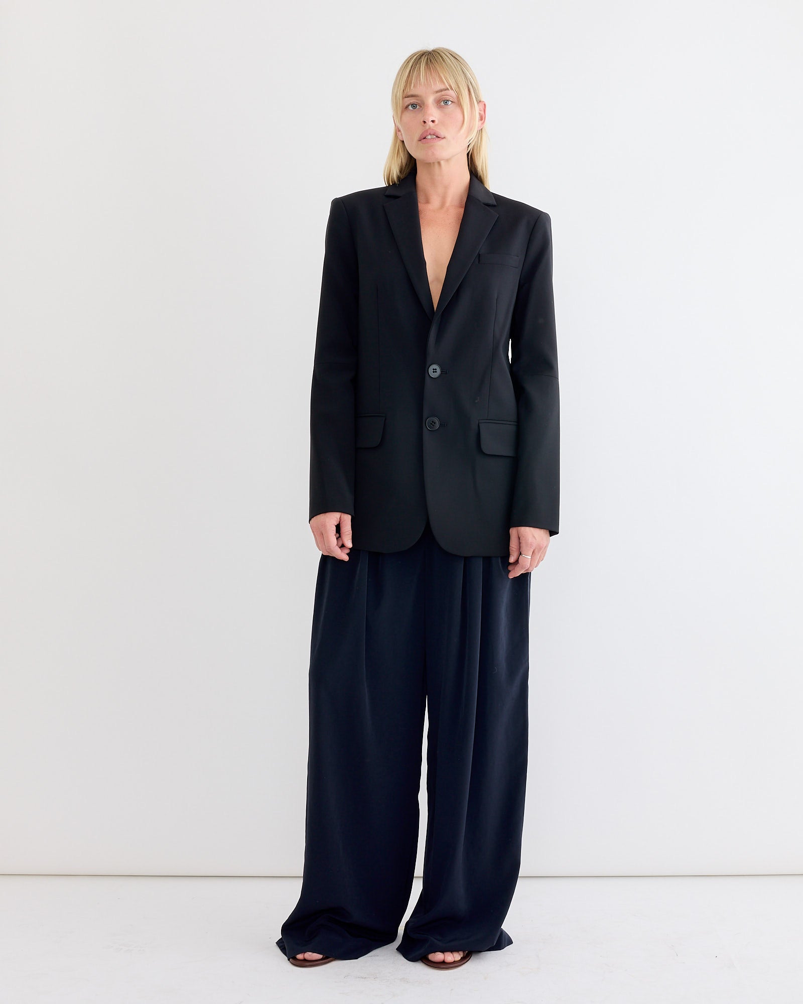 A person with blonde hair stands against a white wall, wearing the Tibi Tropical Wool Cut Out Sleeve Blazer in Black without a visible shirt and loose, wide-leg black pants. Barefoot, they look straight ahead with a neutral expression.