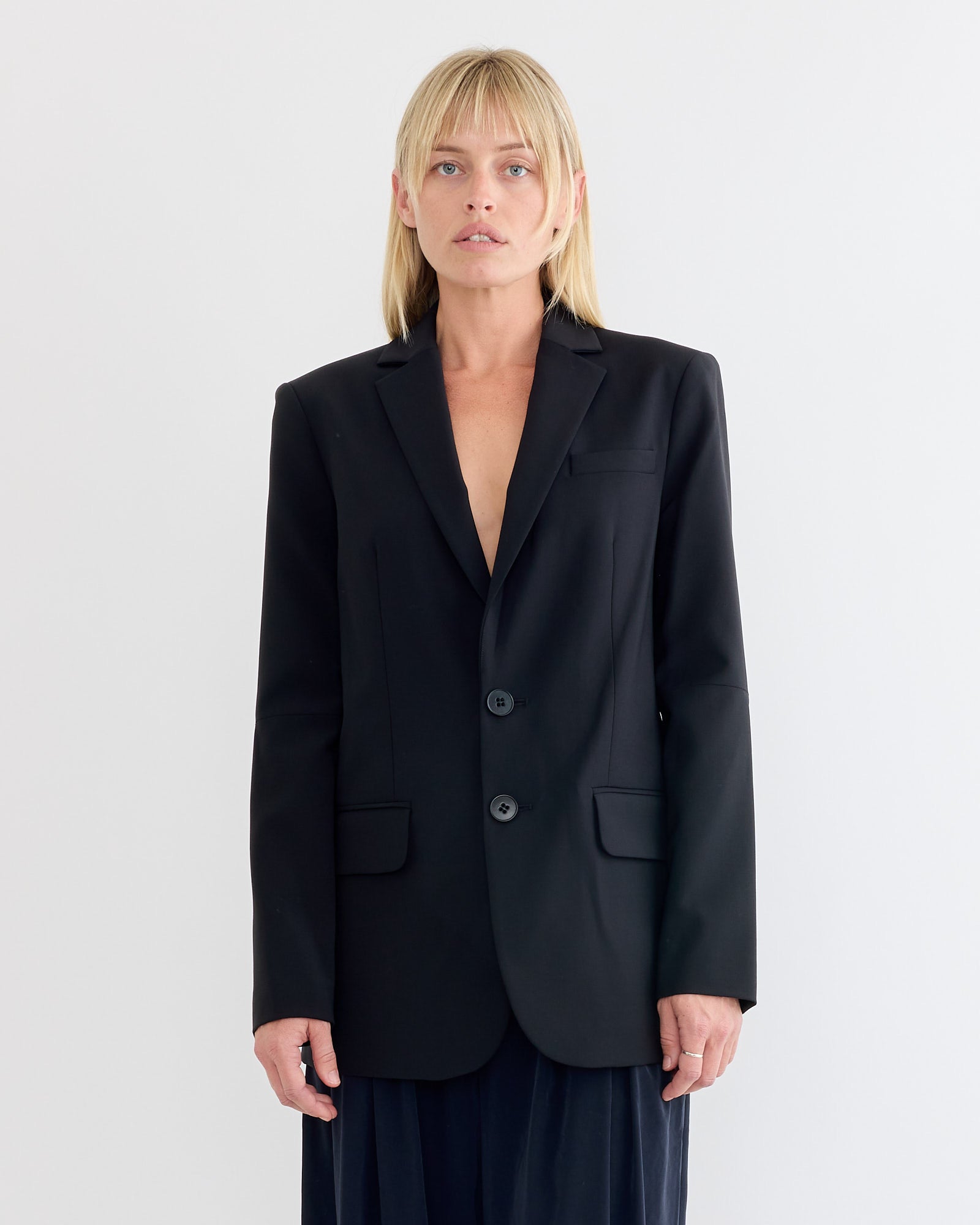 A person with straight blonde hair wears the Tibi Tropical Wool Cut Out Sleeve Blazer in Black, standing against a plain white background and looking directly at the camera.