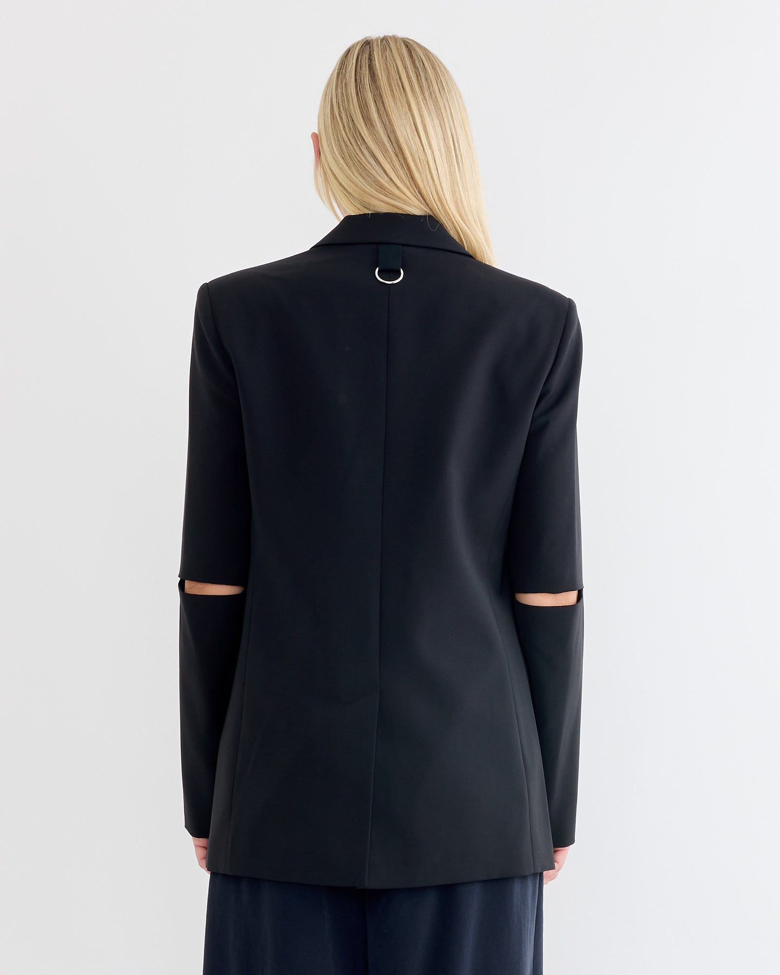 A person with long blonde hair, seen from behind, wears the Tibi Tropical Wool Cut Out Sleeve Blazer in black, featuring elbow cut-outs and a metallic ring at the back of the collar. The background is plain white.